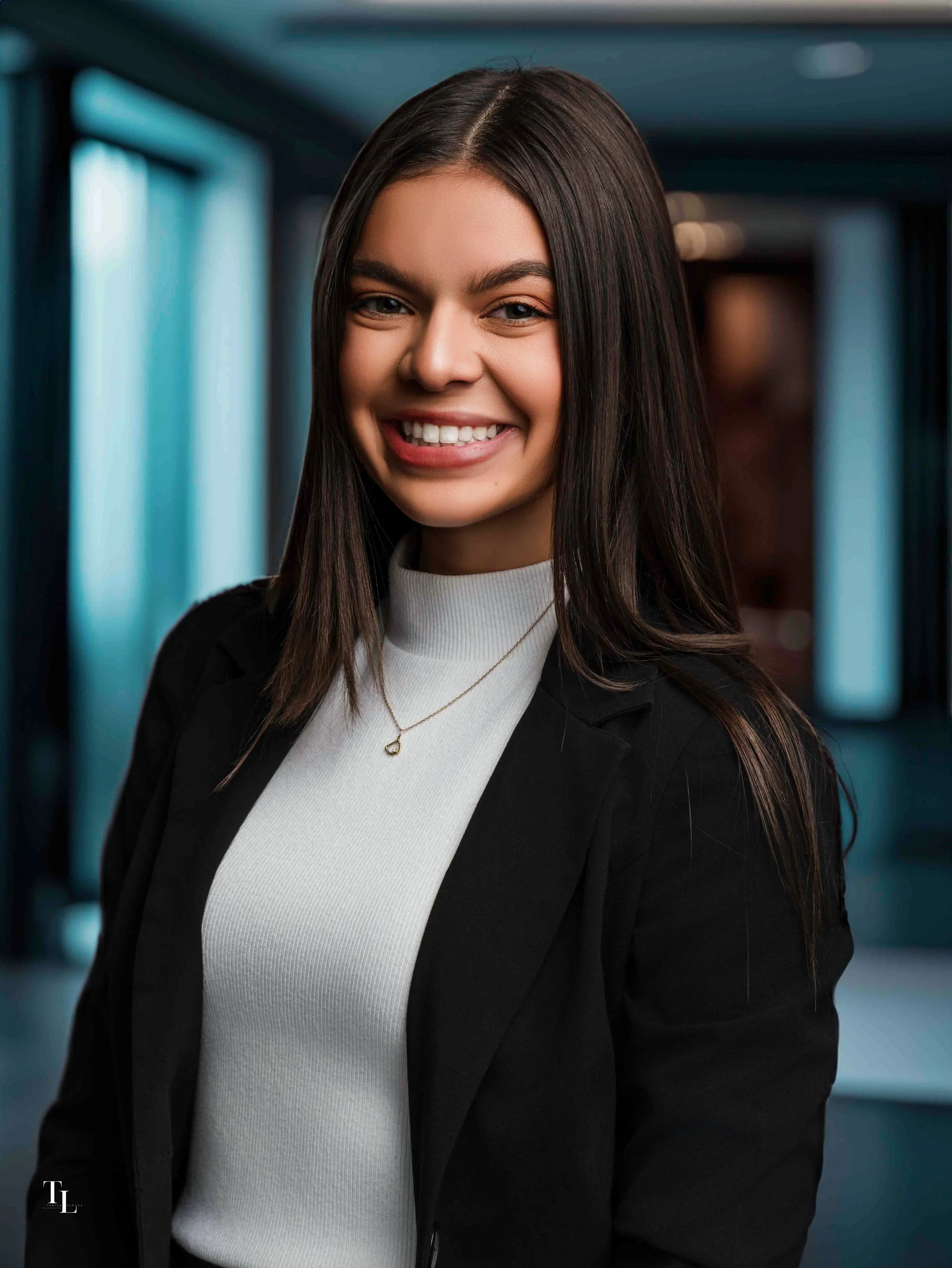 Close-up of a young woman with long dark hair, smiling and wearing a white turtleneck sweater, black blazer, and a delicate necklace, standing indoors with blurred blue-toned background.