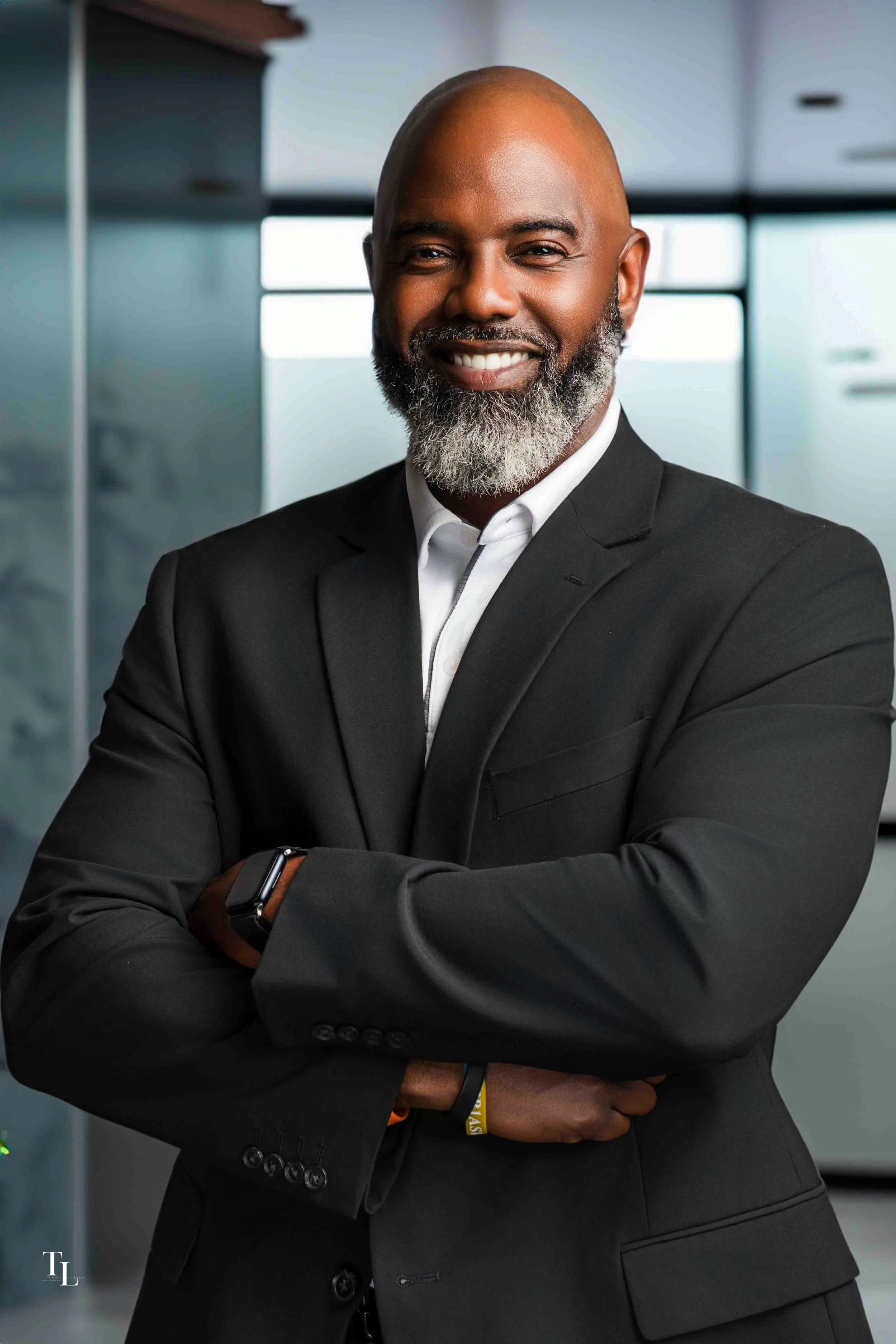 A smiling man with a bald head, gray beard, dressed in a black suit with a white shirt, standing in a modern office setting with large windows.