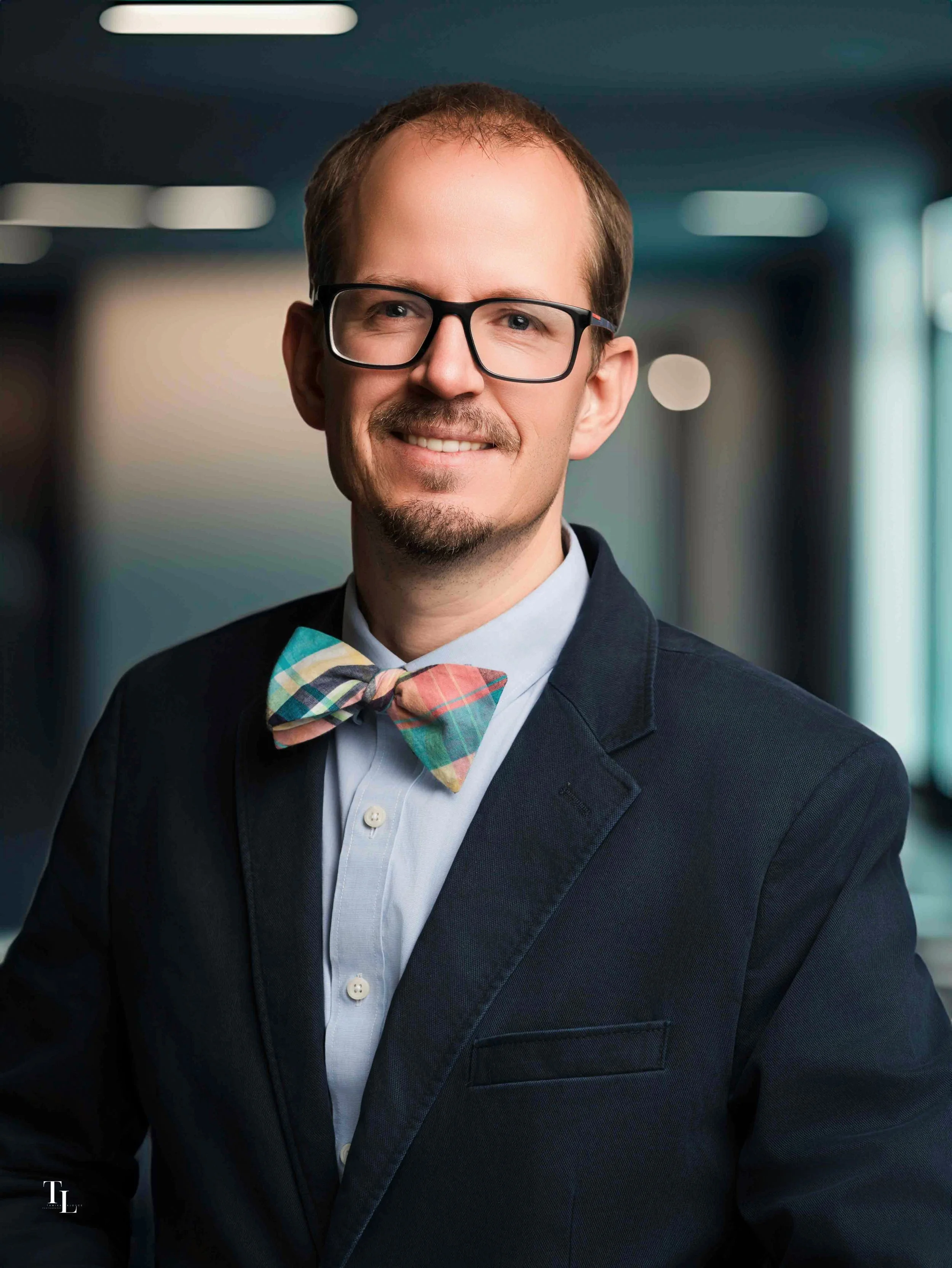 A man with glasses and a mustache wearing a suit and a colorful bowtie, smiling in a professional setting.