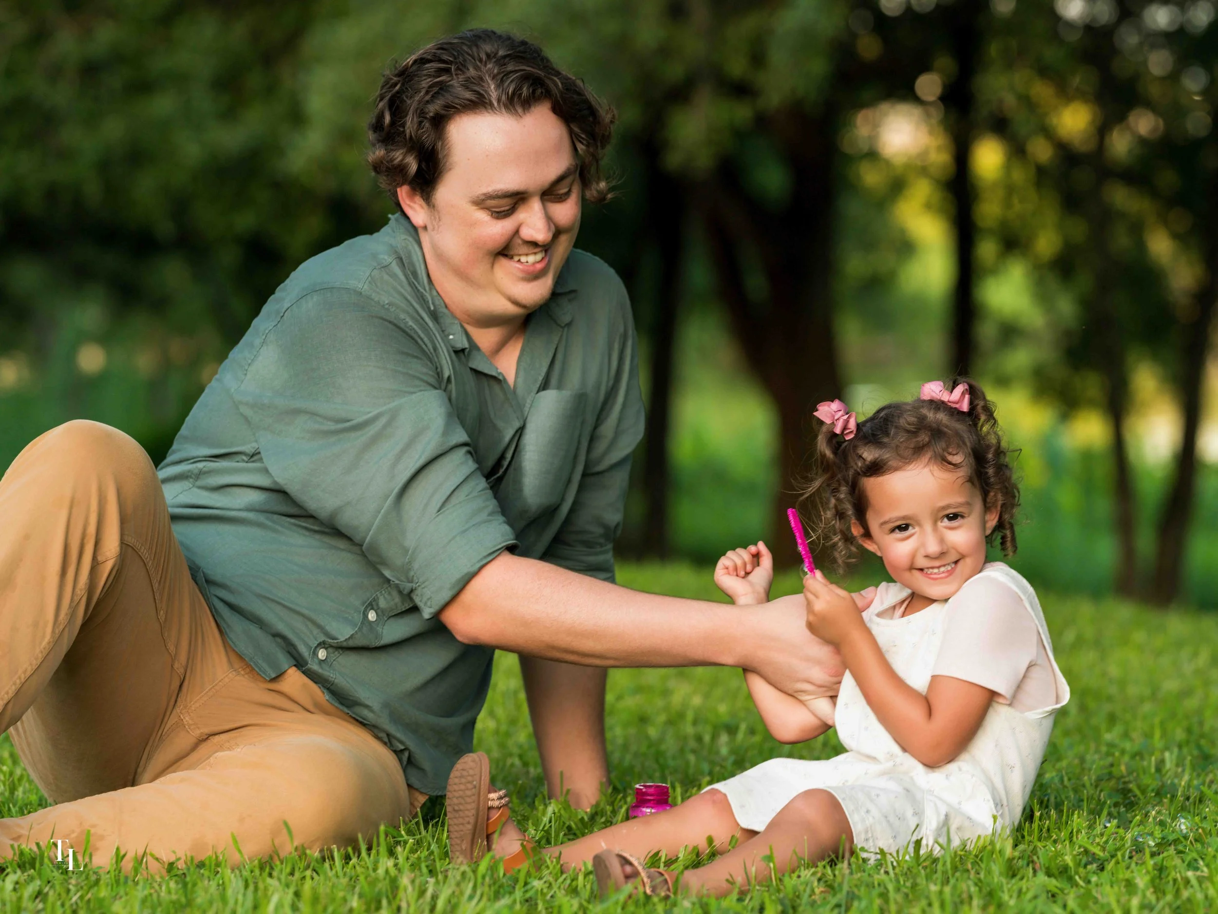 A smiling young man with curly hair and a girl with pigtails sitting on grass in a park, playing together and holding a pink bottle surrounded by trees.