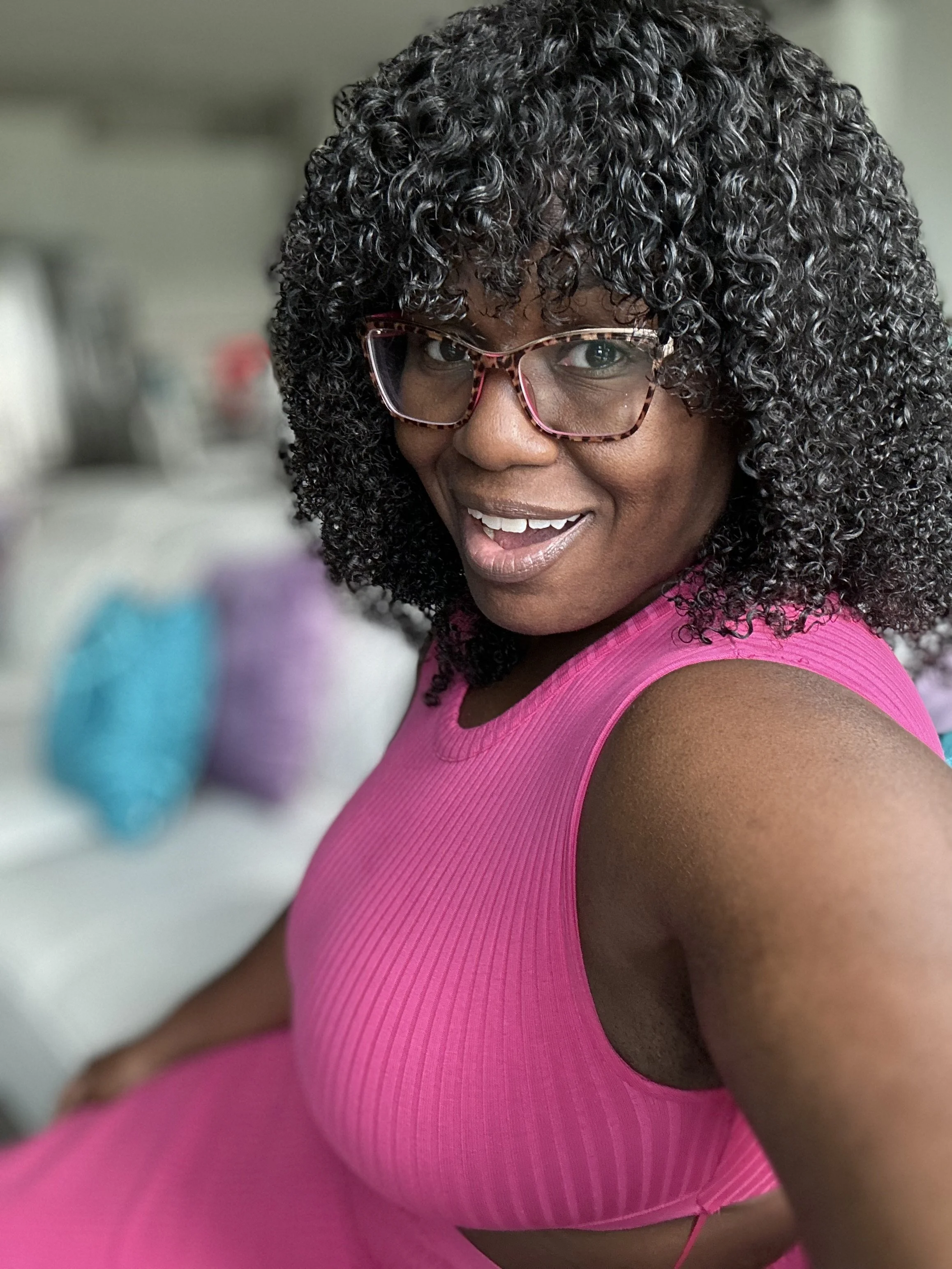 A woman with curly black hair wearing glasses and a pink sleeveless top, smiling at the camera.