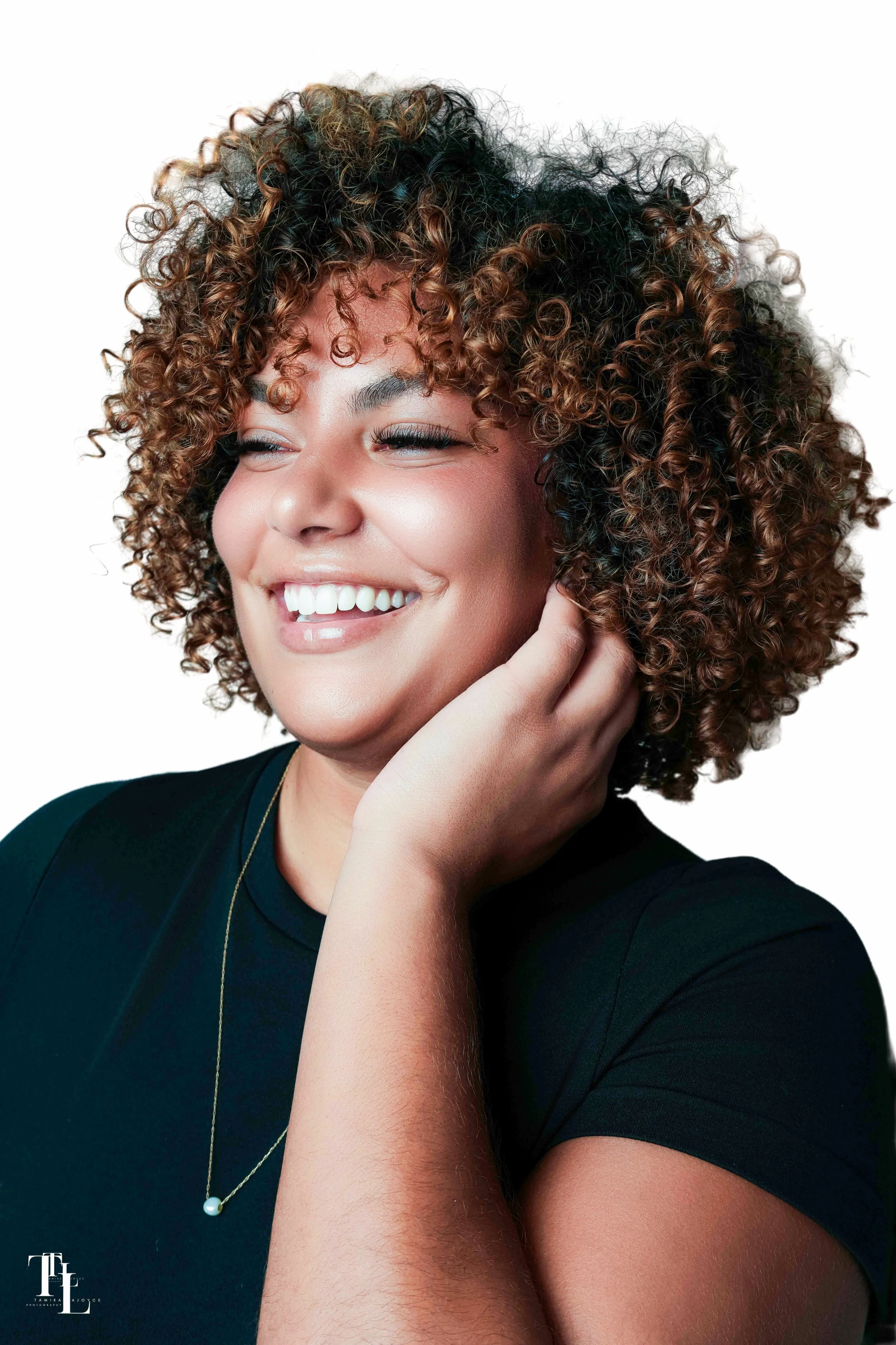 Close-up of a smiling woman with curly hair, wearing a black top, gold necklace with a pearl, and touching her neck.