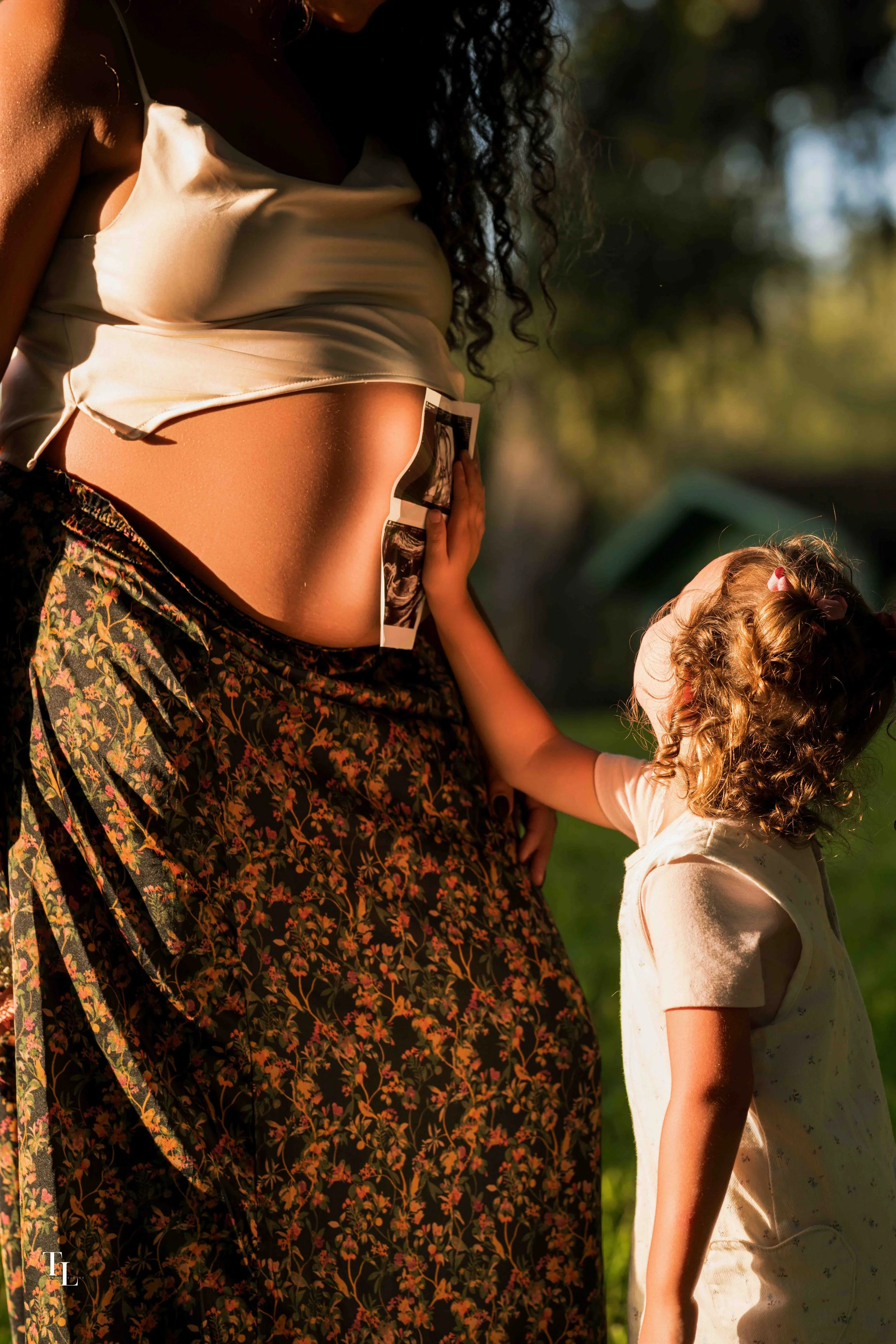 A woman holding ultrasound pictures to a young girl, both outside in sunlight, with a blurred background of trees.
