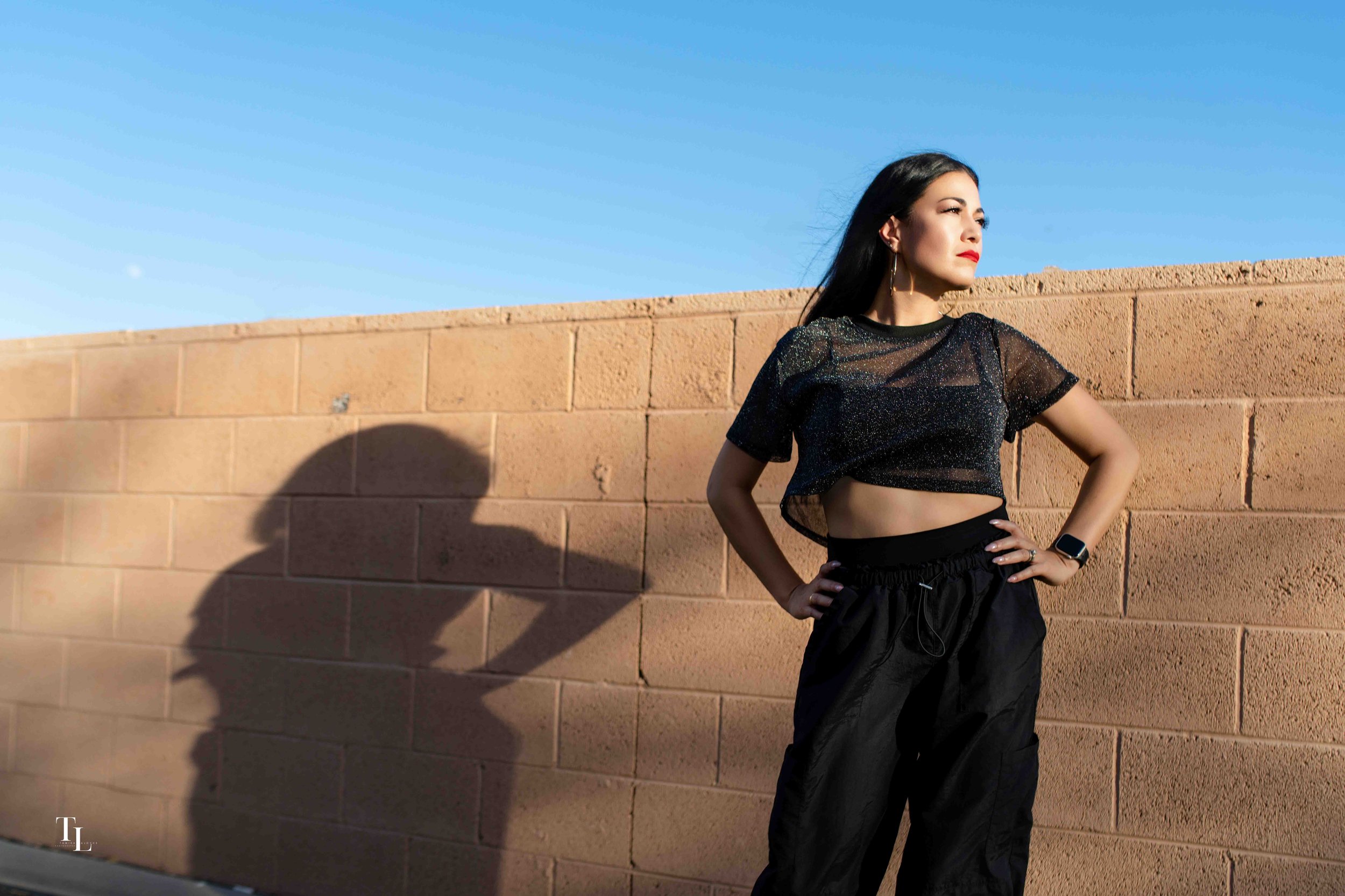 A woman with black hair wearing a black, shiny, short-sleeved top and black pants stands with her hands on her hips, looking to the right, against a brick wall under a clear blue sky.