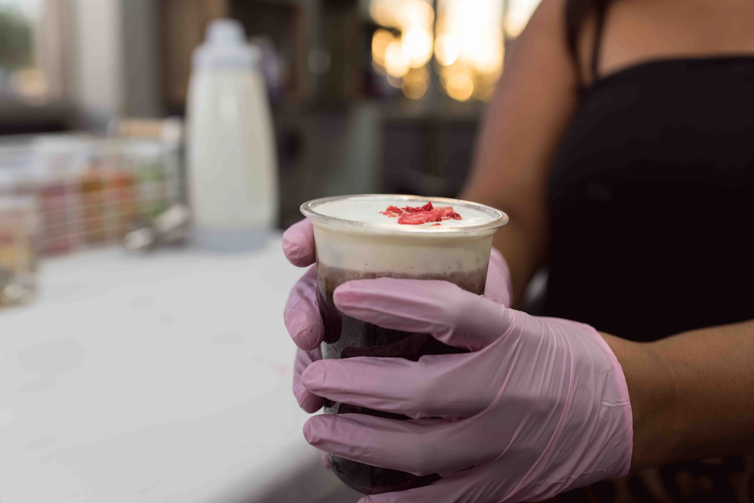 A hand wearing a pink glove holds a small cup of dessert with white topping and pink sprinkles, with a blurred background of a person and indoor lighting.