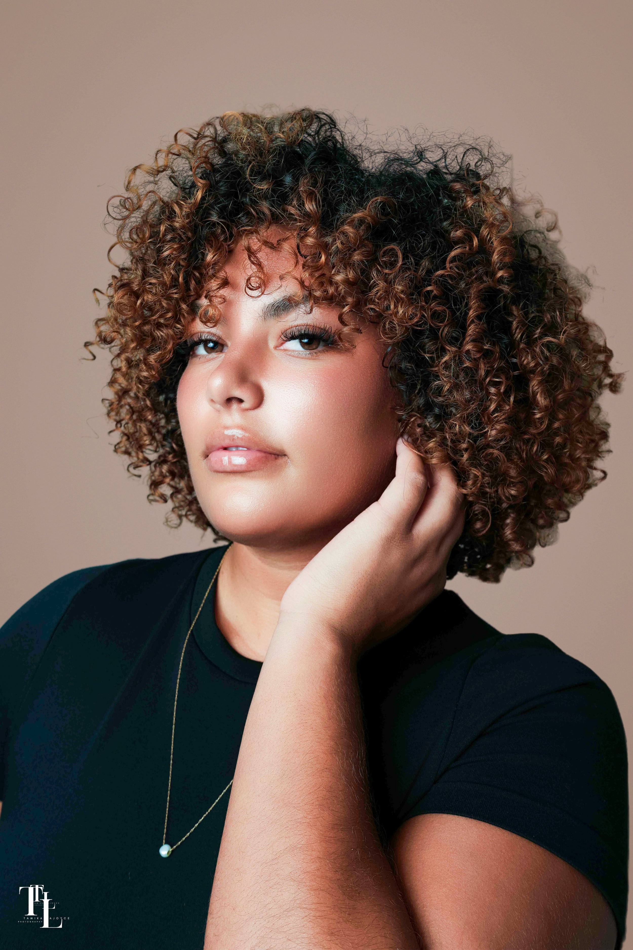 A young woman with curly brown hair, wearing a black shirt and a delicate necklace with a pearl pendant, resting her head on her hand against a plain background.