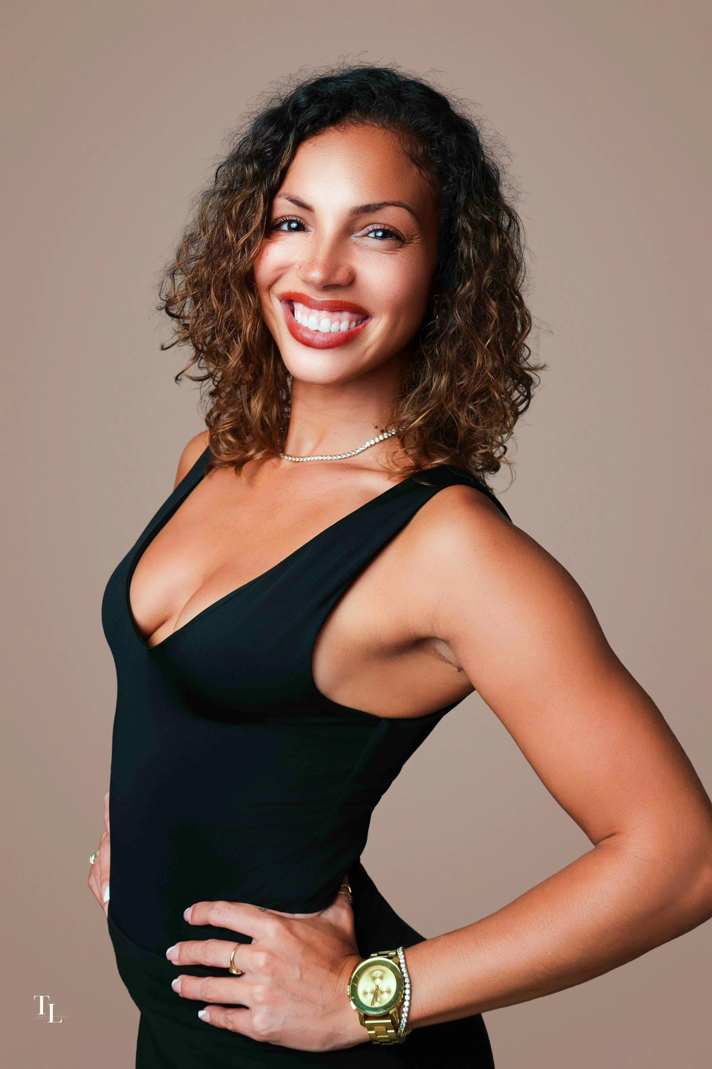Portrait of a woman with shoulder-length curly hair, wearing a black sleeveless dress, smiling, with a gold watch and bracelet, and posing with one hand on her hip against a plain beige background.