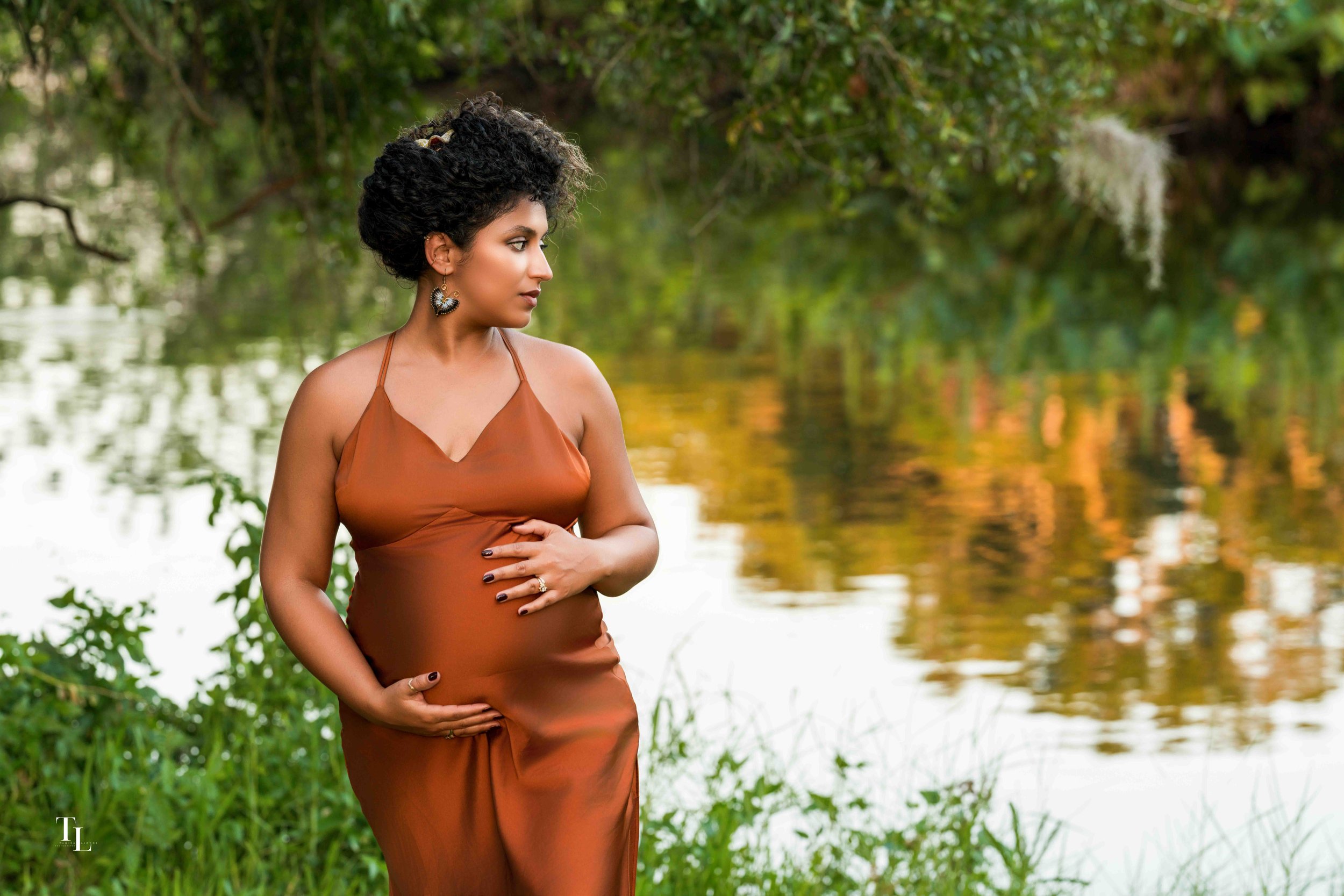 A woman with curly black hair wearing a rust-colored dress stands by a river with green trees and foliage in the background.