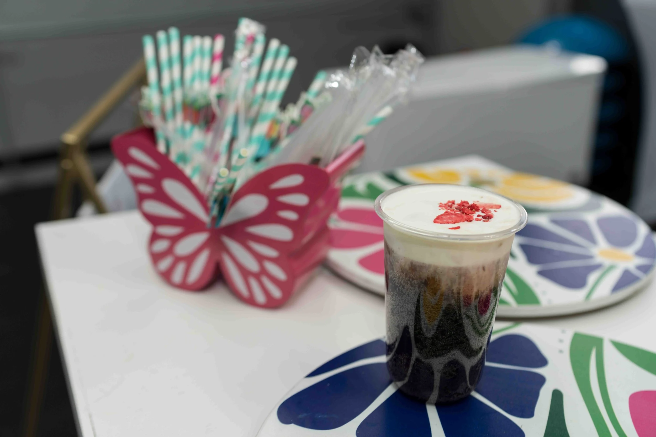 A chocolate milkshake with whipped cream and red sprinkles in a tall glass, placed on a table with a colorful patterned tablecloth. In the background, there is a pink decorative container holding striped paper straws and a plate with yellow slices, p