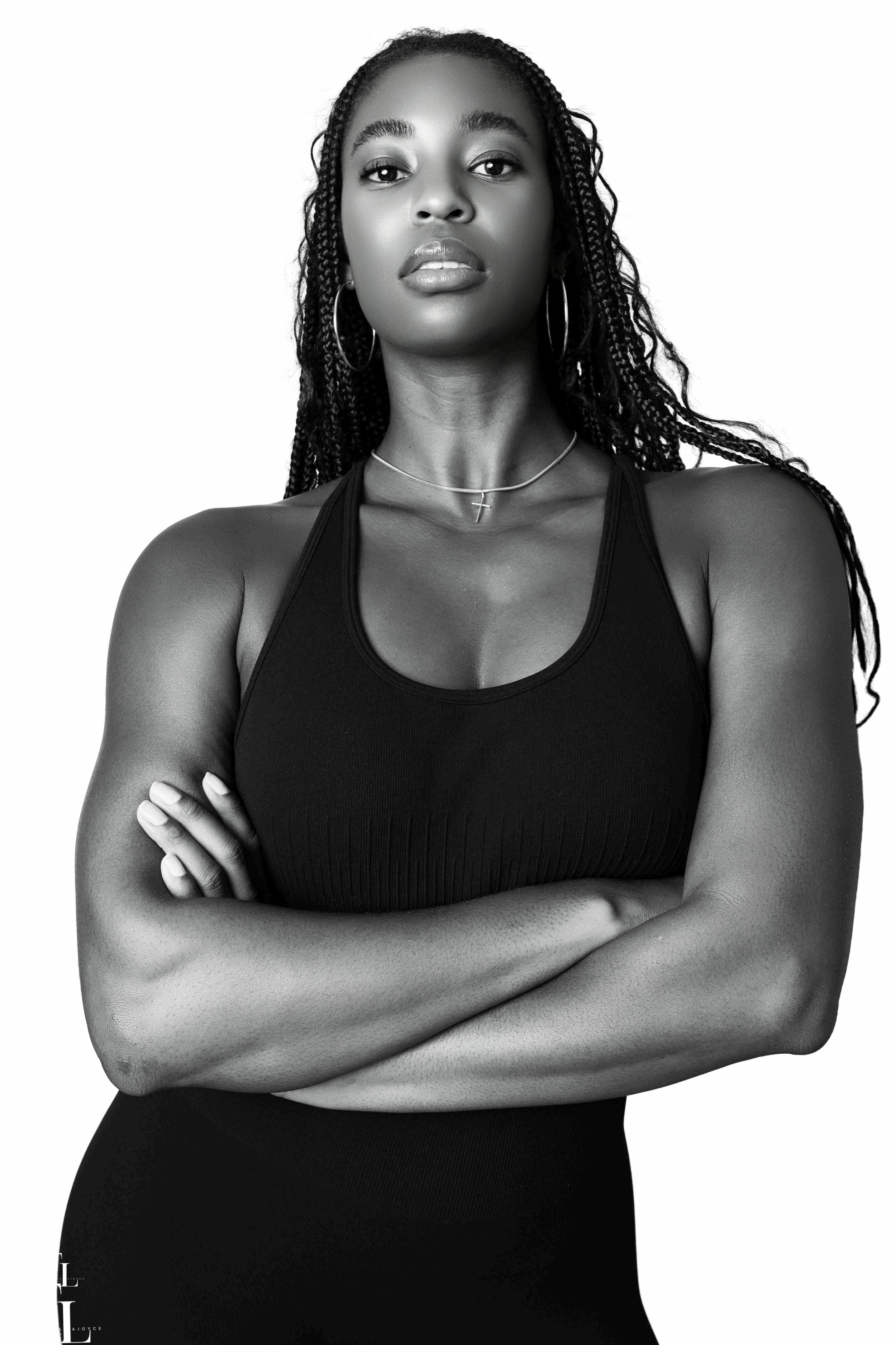 Black and white portrait of a confident woman with braided hair, wearing hoop earrings, a choker necklace with a cross pendant, and a sleeveless top, with her arms crossed.