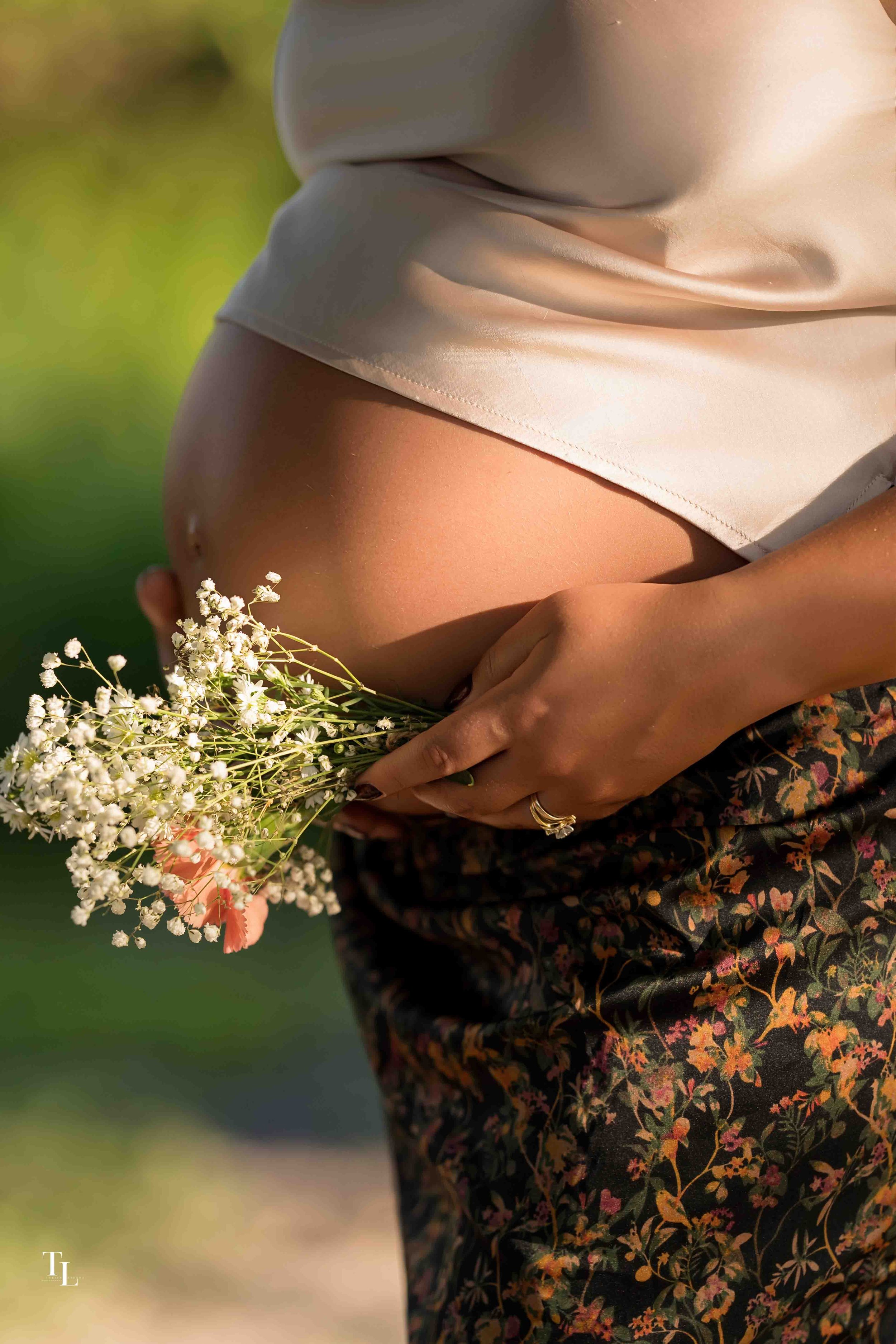 Close-up of a woman wearing a beige satin top, showing her midriff, with her left hand holding a small bouquet of white baby's breath flowers, and dressed in a floral skirt with orange and pink flowers. The background is blurred outdoors with greener