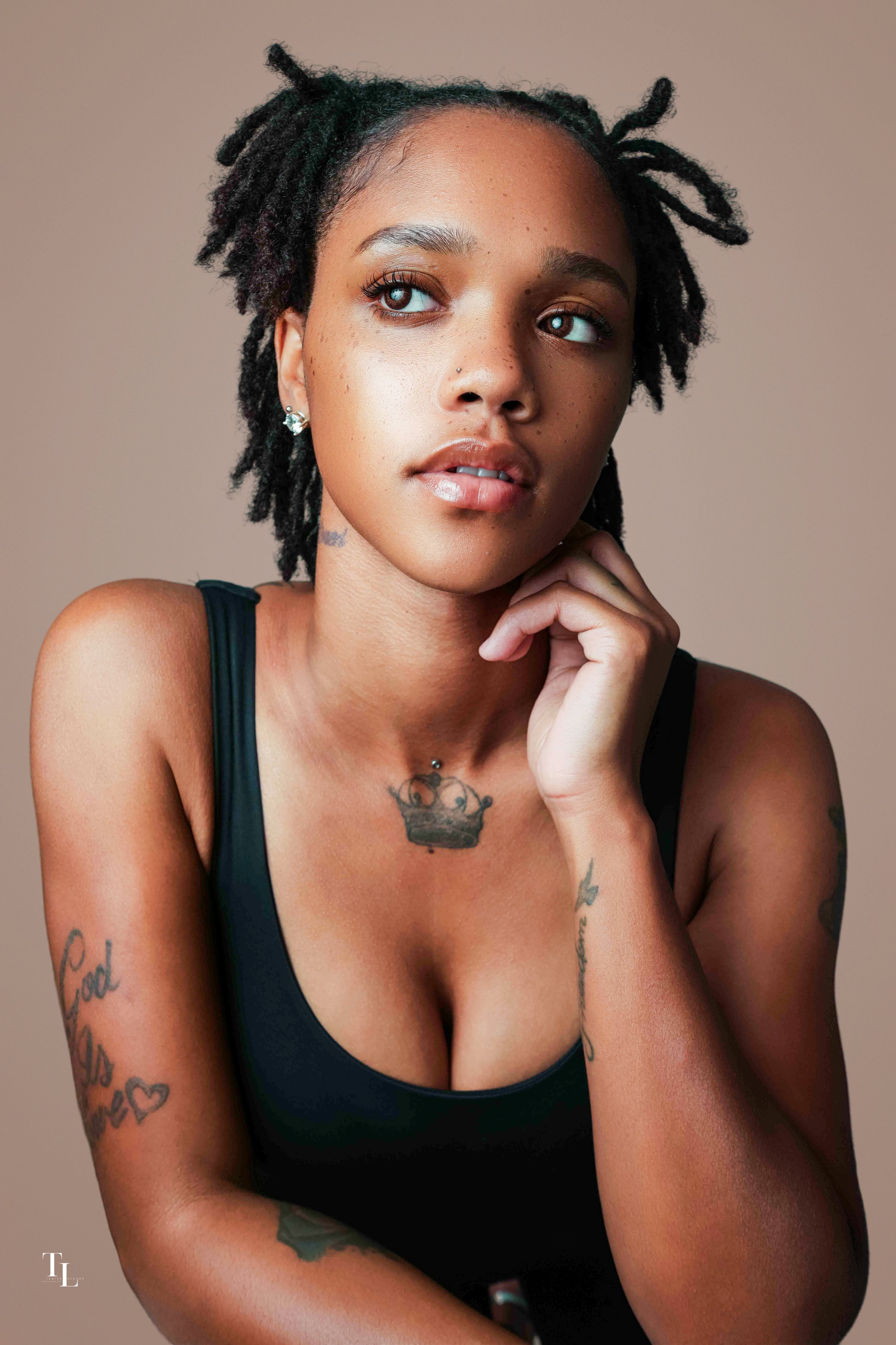A young woman with short dreadlocks and multiple tattoos, resting her chin on her hand, wearing a black tank top, against a neutral background.