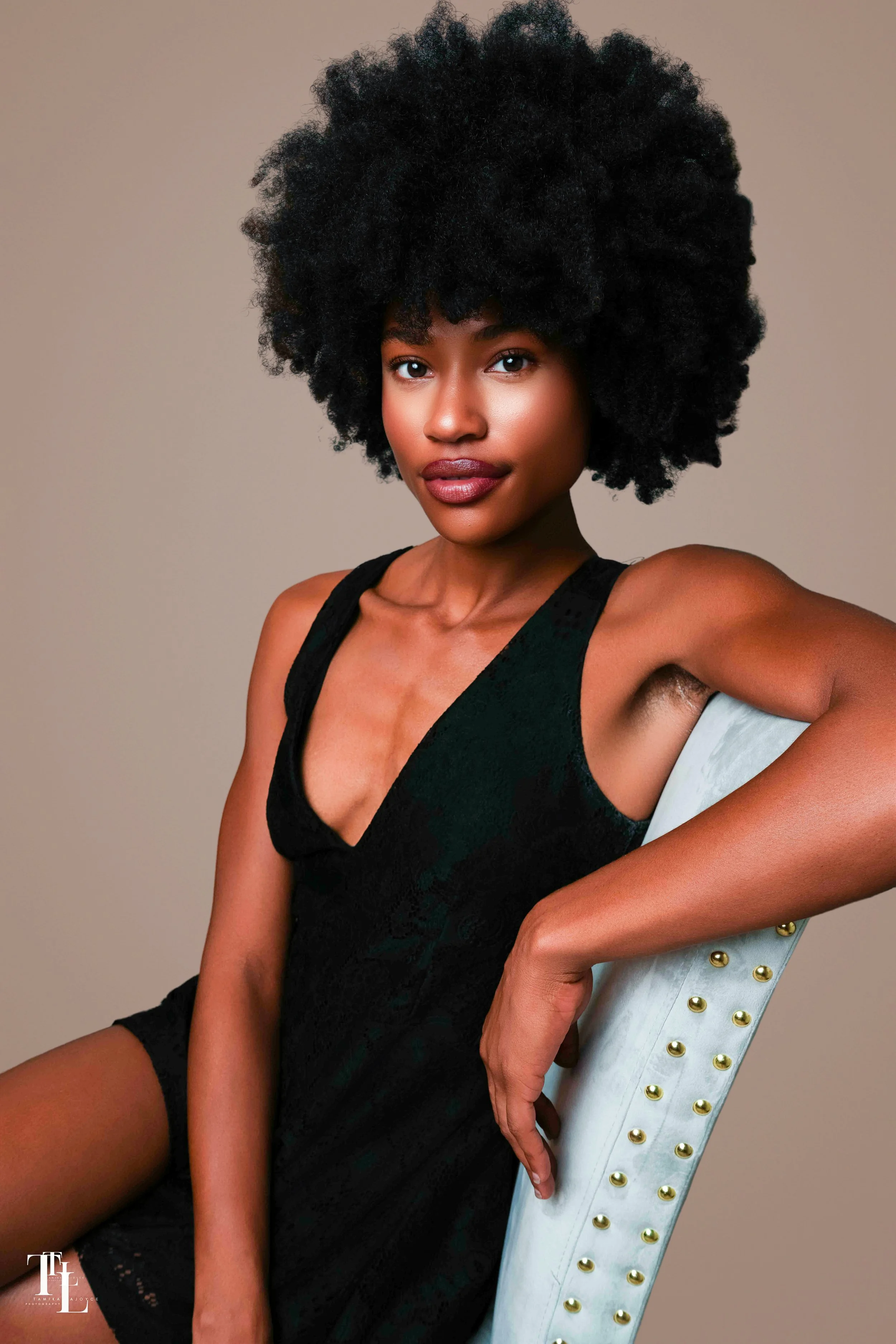 Portrait of a young woman with dark skin, wearing a black sleeveless dress, sitting on a white chair with gold stud accents, against a plain background.
