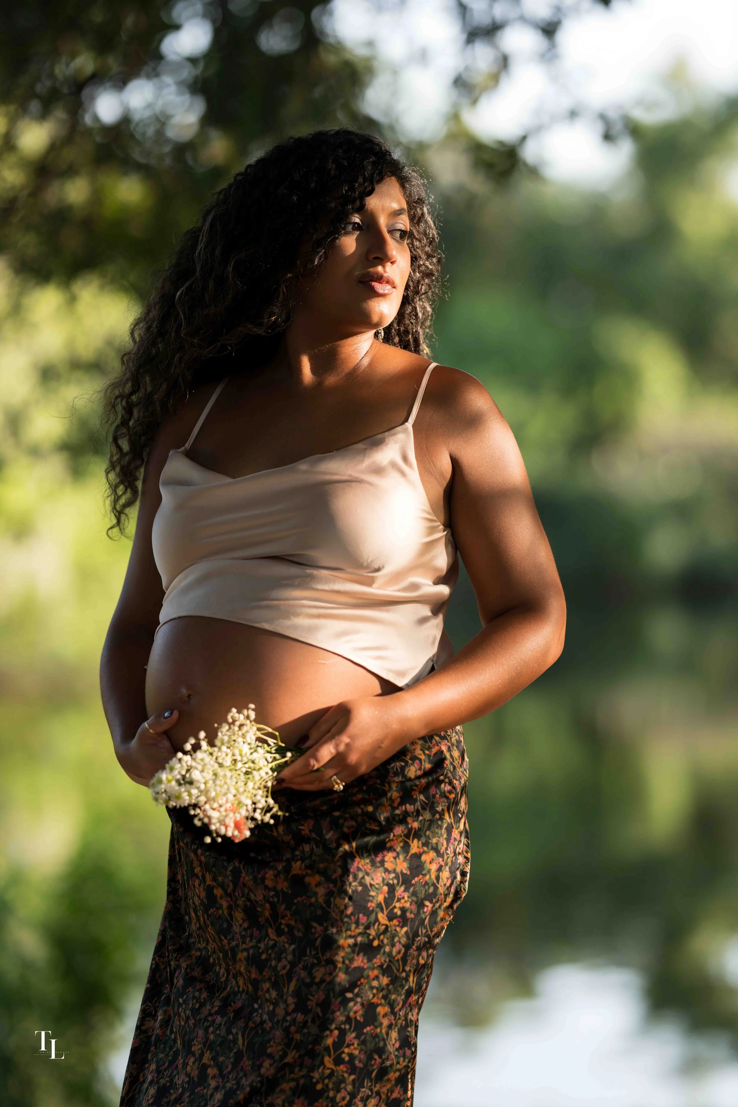A pregnant woman with curly hair standing outdoors in a natural setting with blurred green foliage in the background. She is wearing a beige satin top with thin straps and a patterned skirt, holding a small bouquet of white flowers.