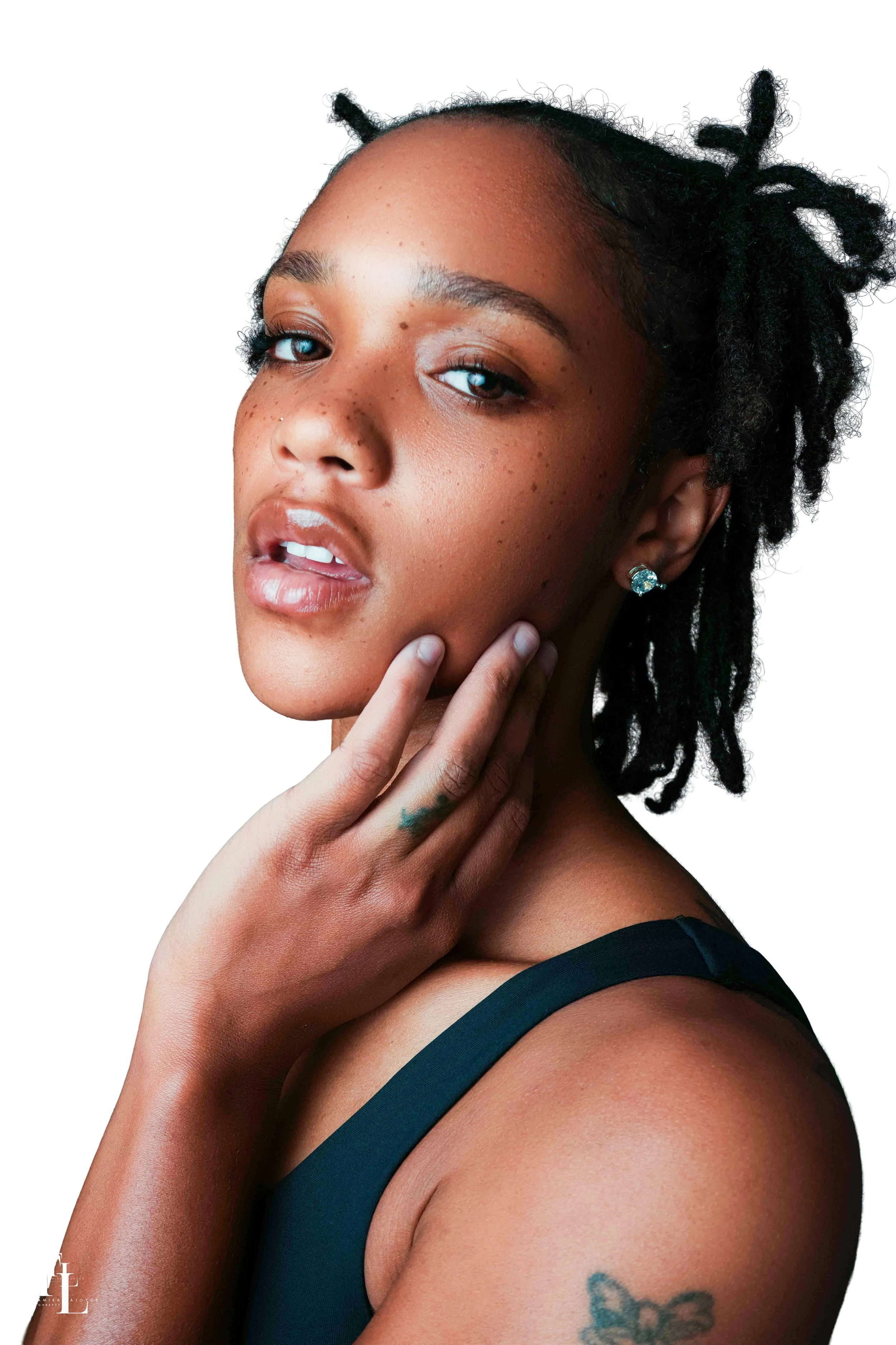 A woman with dark freckled skin and dreadlocks posed against a white background, wearing a black tank top and earrings.