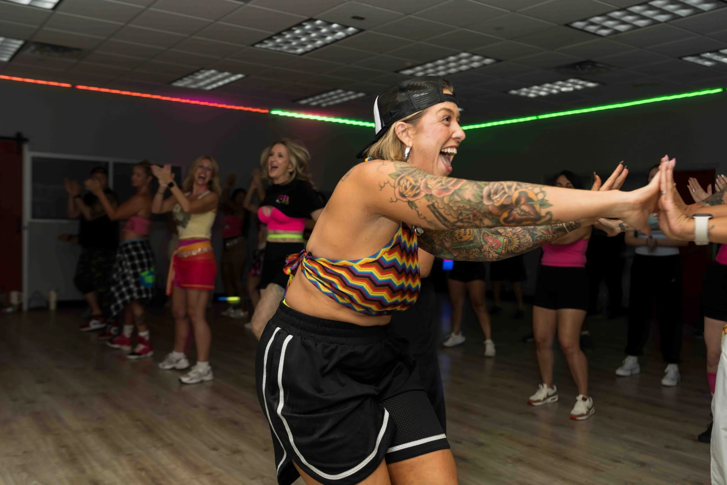 A group of women dance and laugh in a room with black walls and wooden flooring, illuminated by red and green laser lights on the ceiling. The women are dressed in colorful, casual dance attire.