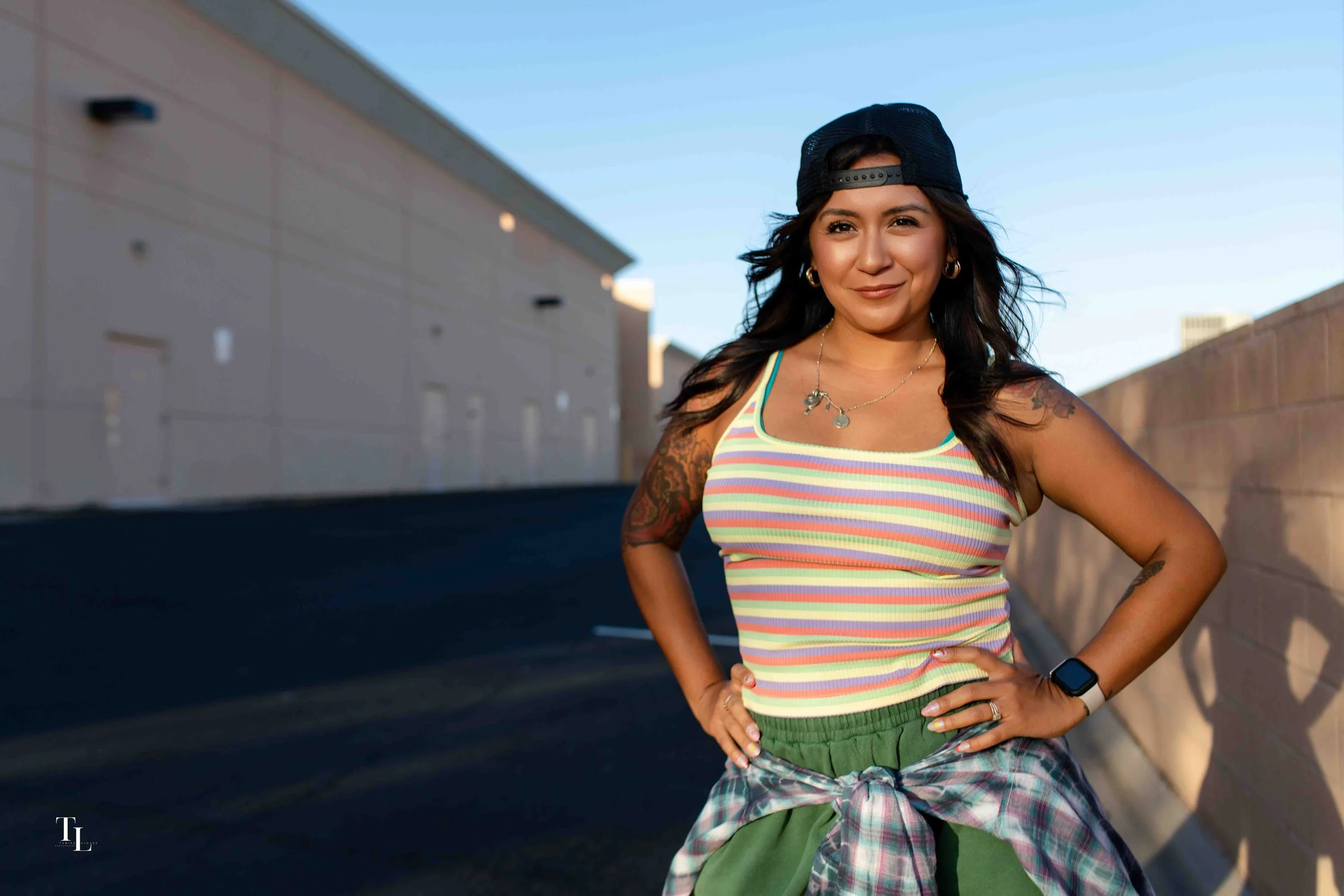 A woman wearing a black backwards cap, gold hoop earrings, a colorful striped tank top, and green pants stands outdoors with a confident pose, smiling, with a beige wall and blue sky in the background.