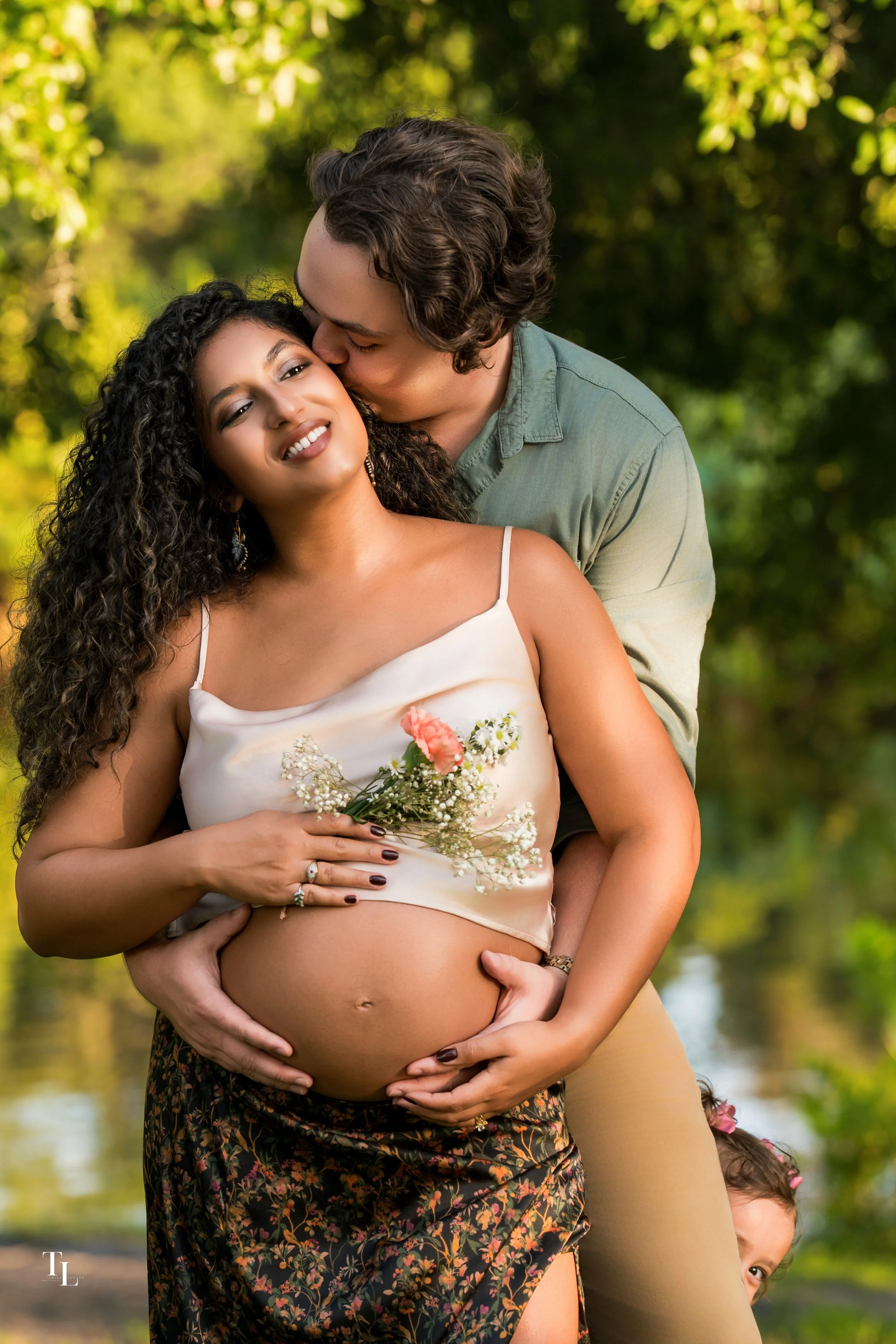 A pregnant woman holding a flower bouquet and smiling, with a man behind her kissing her forehead, outdoors with green trees in the background.