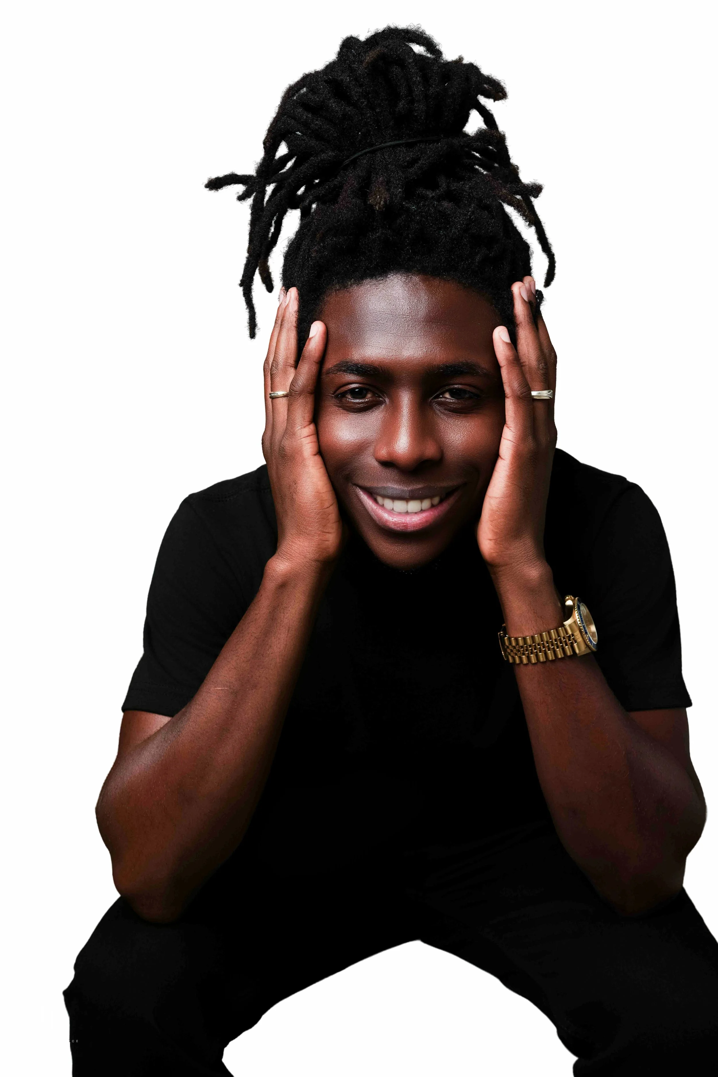 A smiling person with dreadlocks tied up, wearing a gold watch, rings, and a black shirt, holding their face with both hands, against a white background.