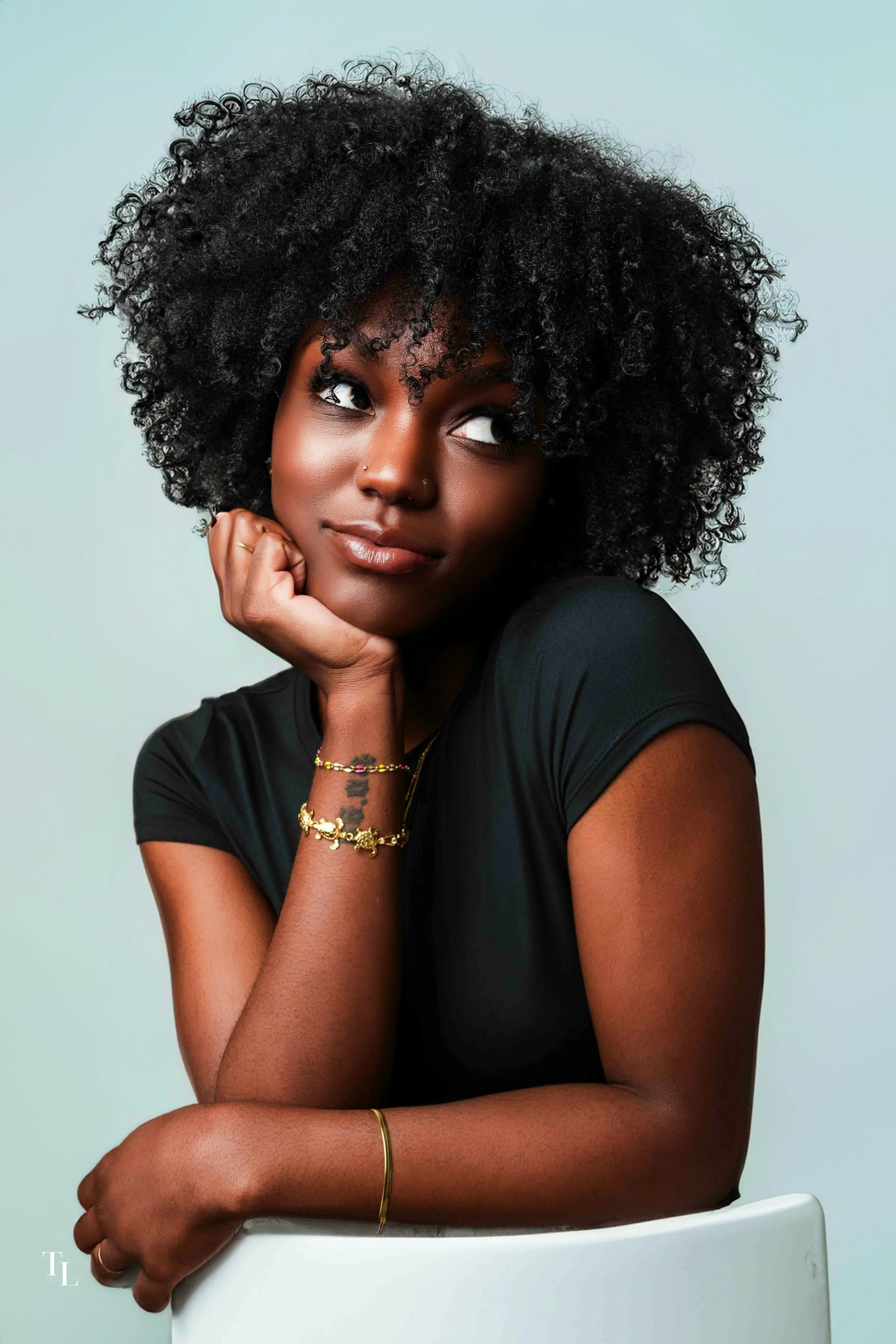 Portrait of a woman with natural curly hair, wearing a black top and gold jewelry, resting her chin on her hand, against a light blue background.