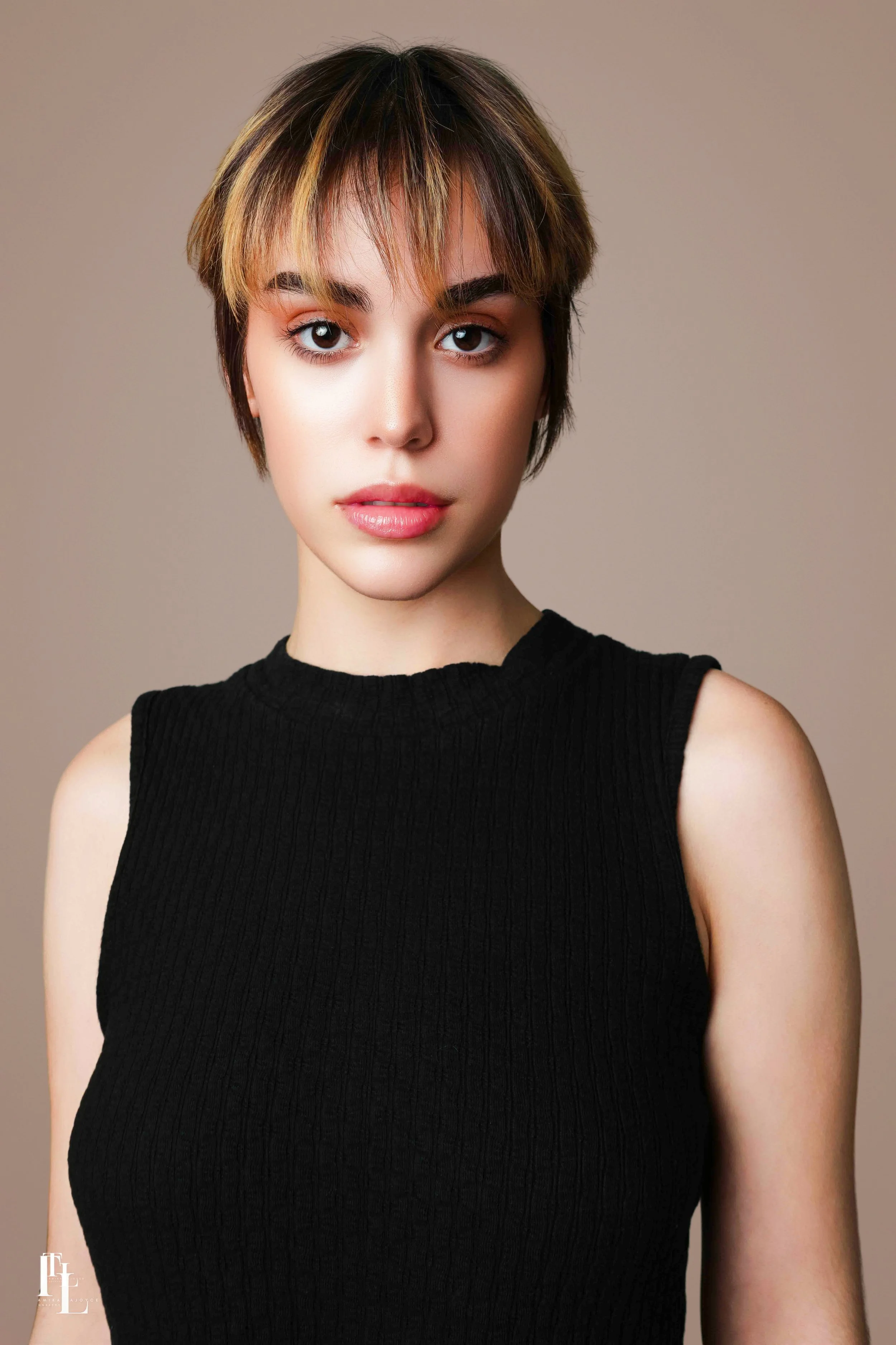 A young woman with short, layered brown hair with blonde highlights, wearing a black sleeveless top, against a plain beige background.