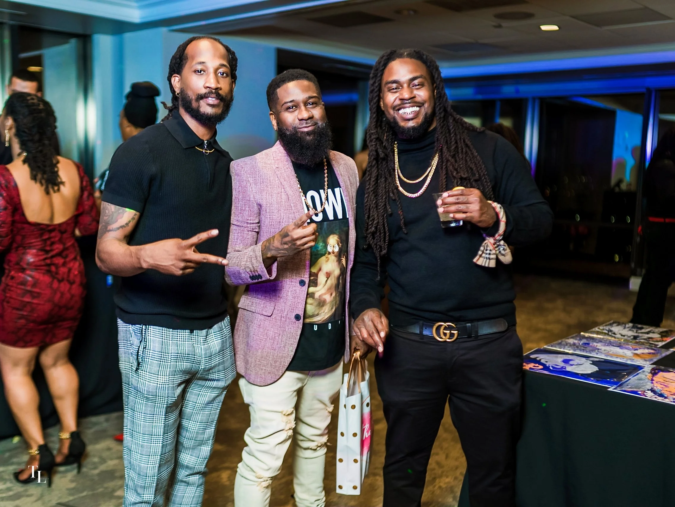 Three men posing at a social event, standing in front of a table with art prints on display. They are smiling and making gestures, with other guests in the background.