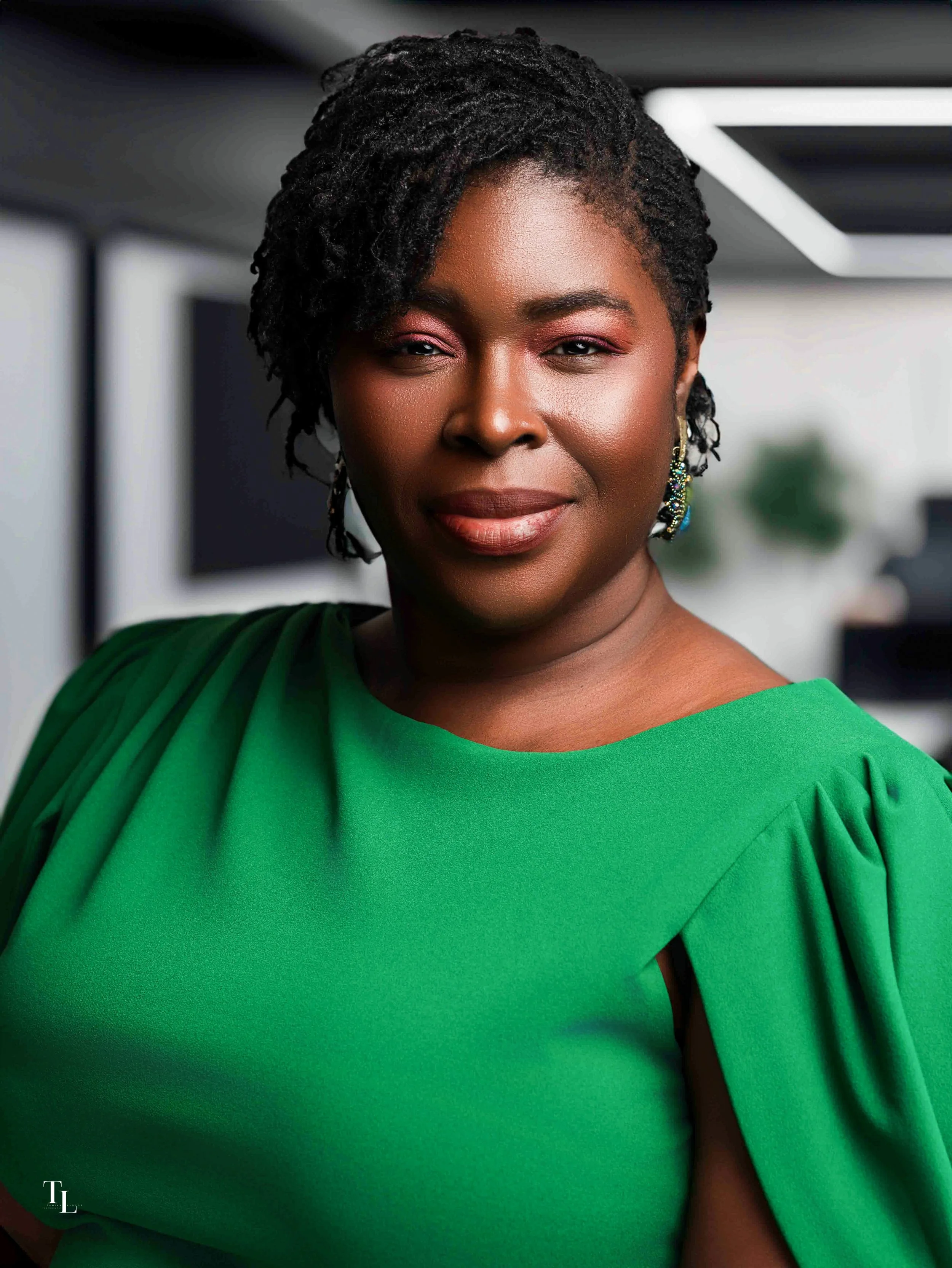 Close-up of a woman with dark skin and styled black hair, wearing a green dress with puffed sleeves and colorful earrings, in an indoor setting with a modern background.