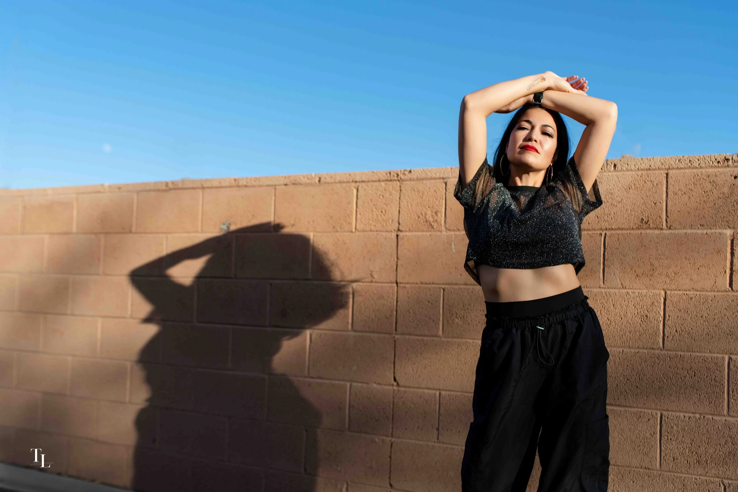 A woman with dark hair and red lipstick standing against a brick wall on a sunny day, with her eyes slightly closed and arms raised above her head.