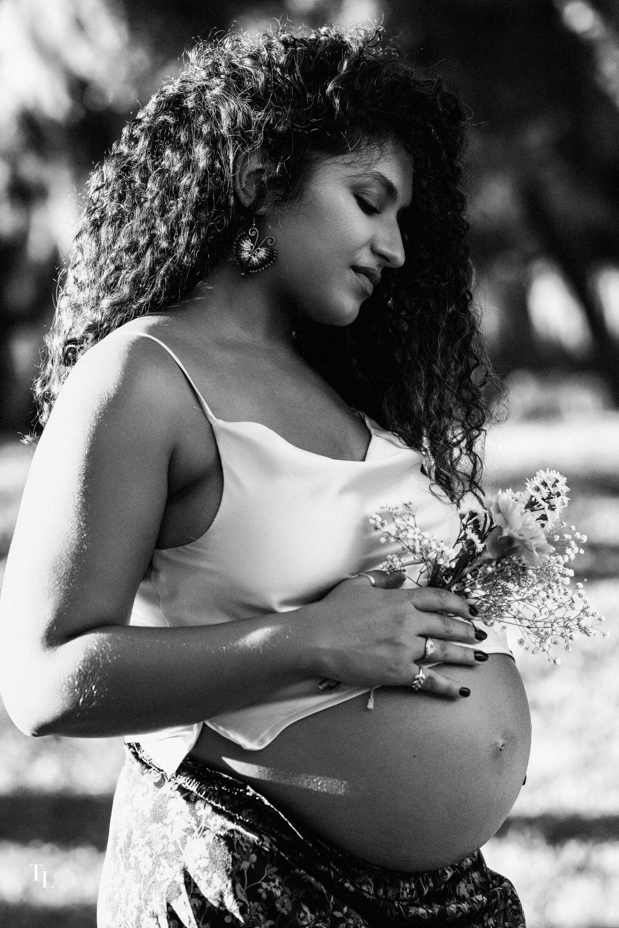 A pregnant woman with curly hair wearing earrings and a sleeveless top, holding a bouquet of flowers on her belly, standing outdoors in a natural setting.