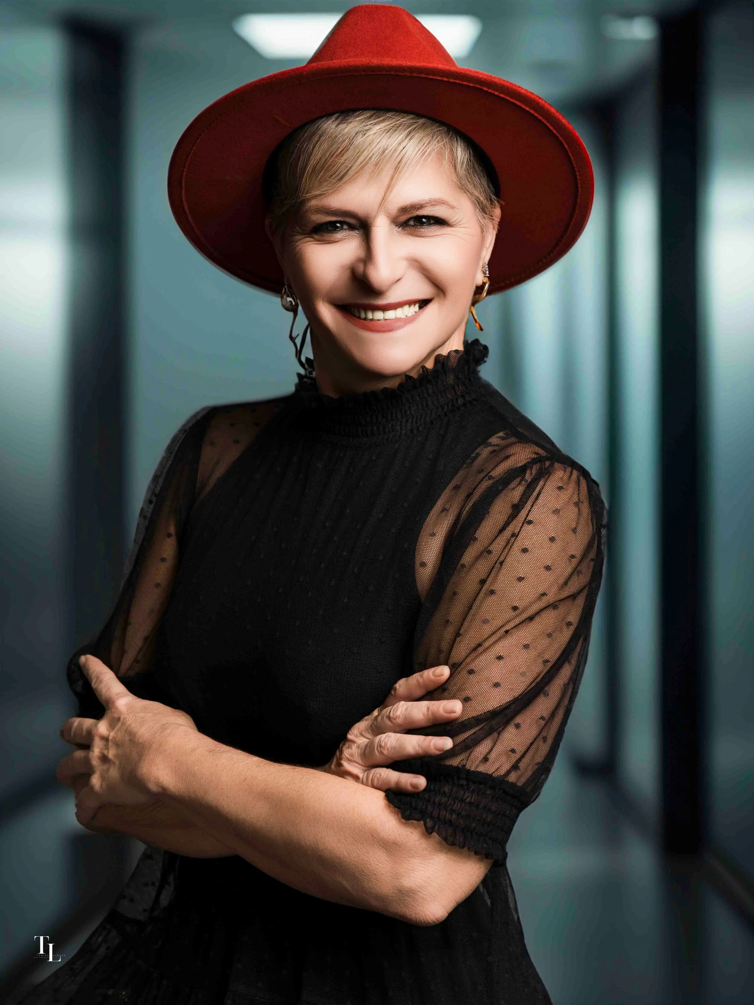 A woman with short blonde hair smiling, wearing a red wide-brimmed hat and a black sheer top with polka dots, crossed arms, standing in a modern, indoor setting.