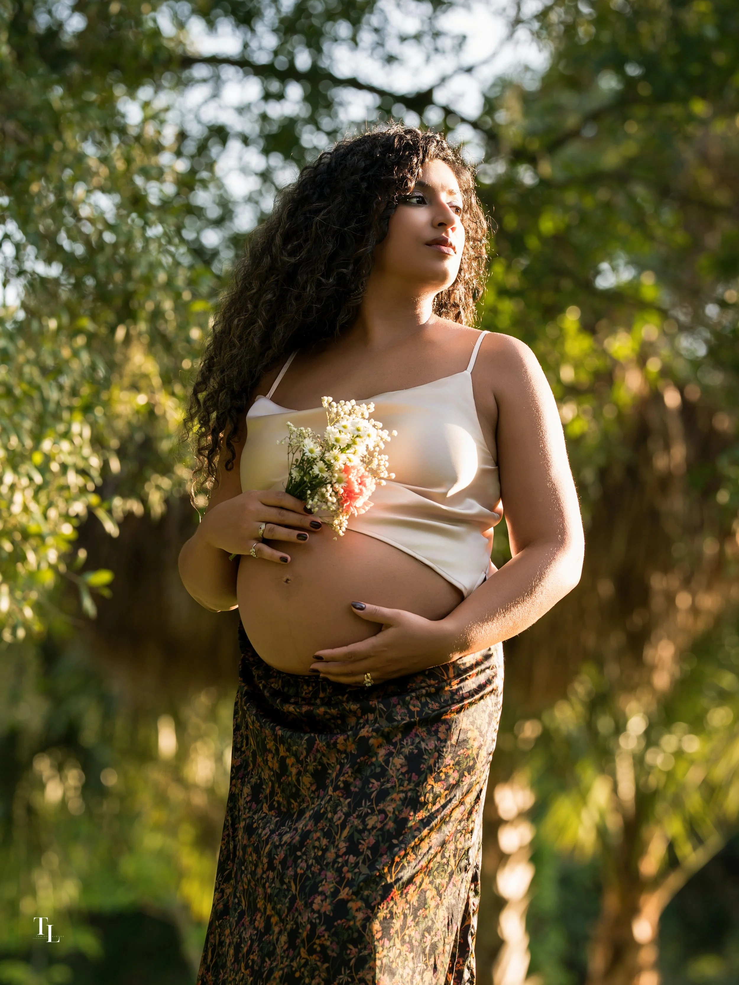 A pregnant woman with long, curly hair stands outdoors surrounded by sunlight and greenery, holding a small bouquet of flowers and gently cradling her belly.
