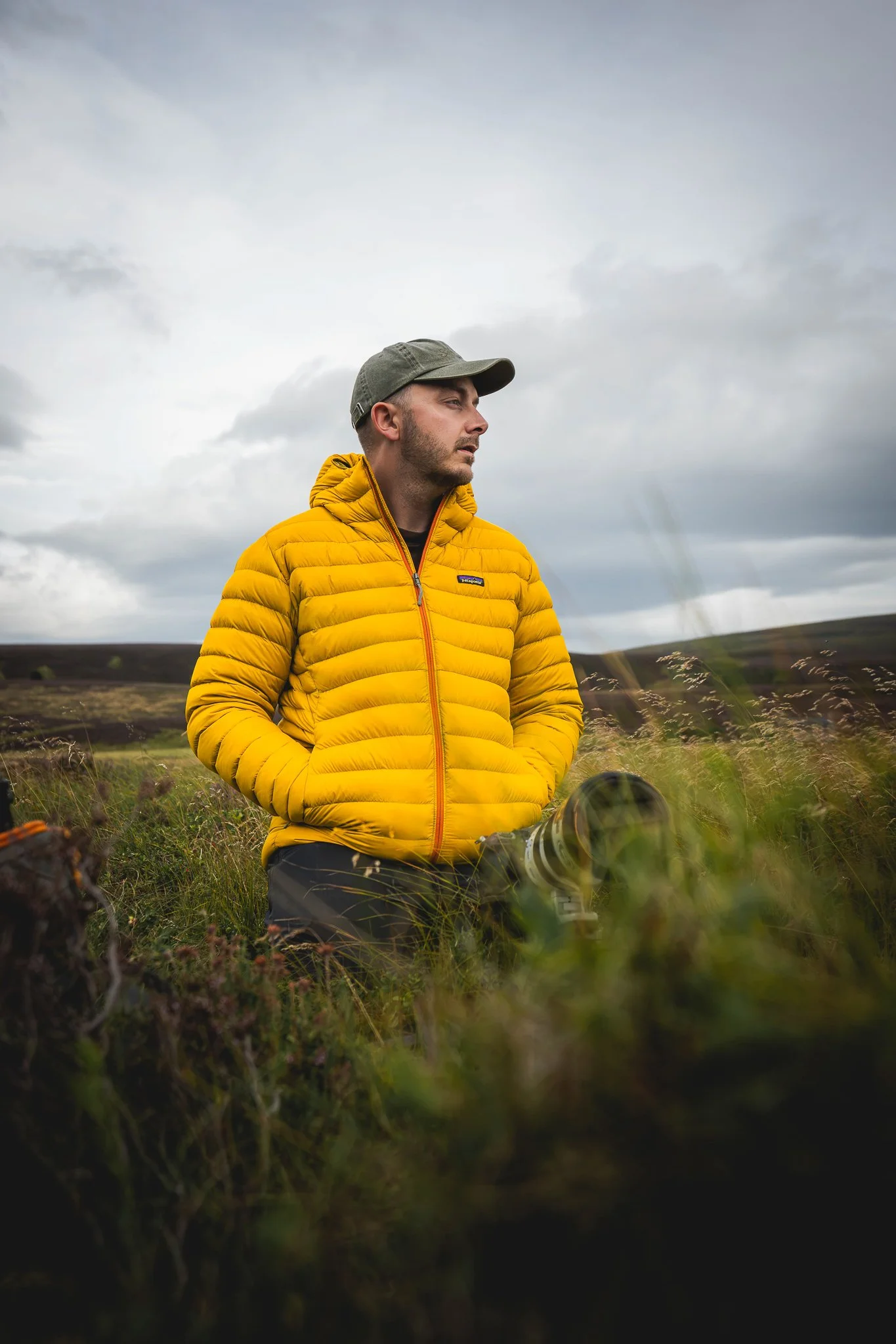 A man in a bright yellow puffy jacket and a gray baseball cap sitting outdoors in a grassy field, with hills and a cloudy sky in the background, holding a camera with a telephoto lens.