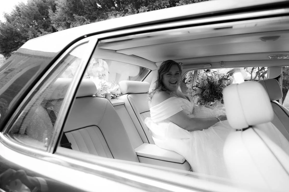 A woman in a wedding dress sitting inside a vintage car, smiling and holding a bouquet of flowers, dorset devon cinematic documentary style wedding photographer