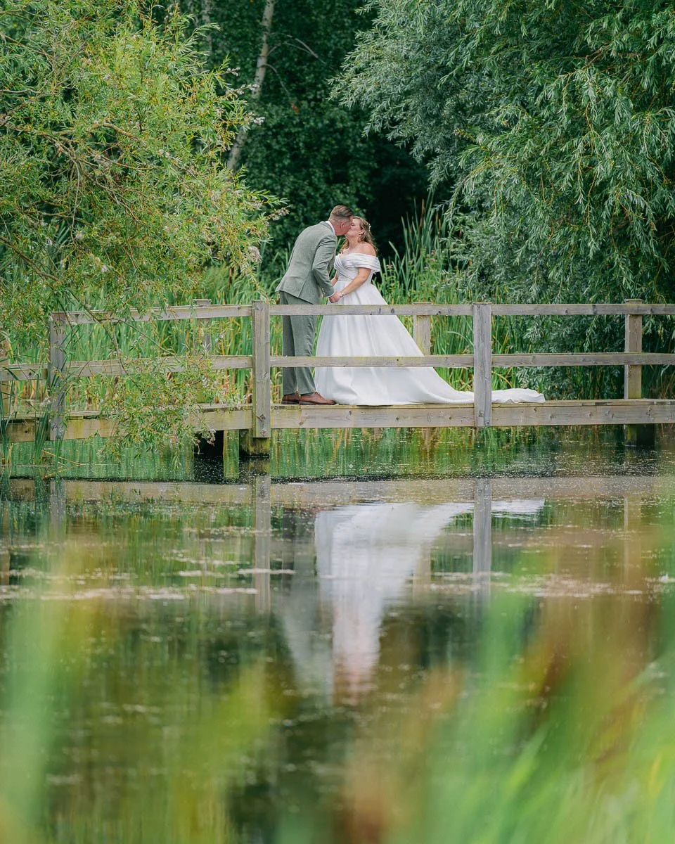 A newlywed couple sharing a kiss on a wooden bridge over a pond, surrounded by lush green trees and foliage dorset devon cinematic documentary style wedding photographer