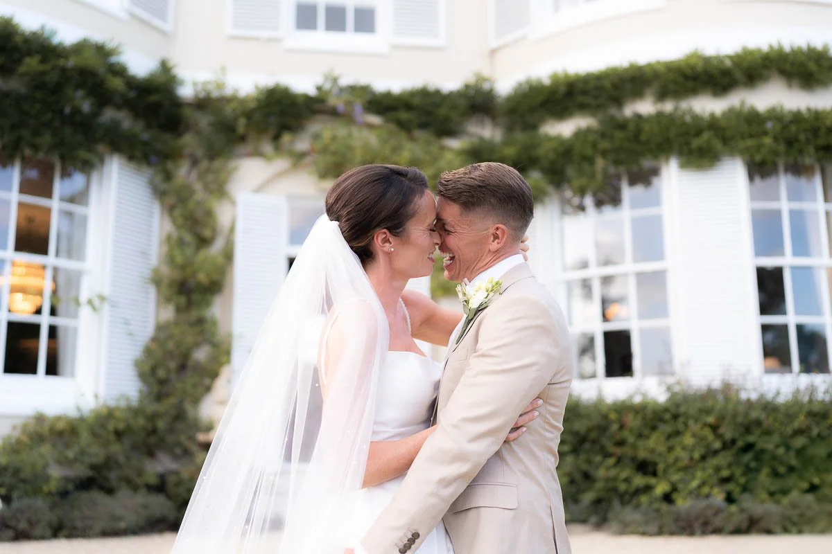 A bride and groom are smiling and touching foreheads, embracing each other outdoors in front of a white building with large windows and greenery dorset devon cinematic documentary style wedding photographer