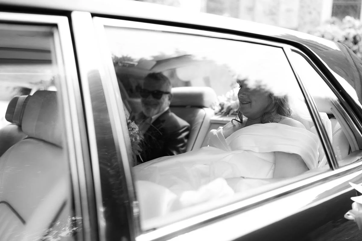 A woman in a wedding dress smiling while sitting in the back of a car, with her dad in sunglasses sitting beside her dorset devon cinematic documentary style wedding photographer