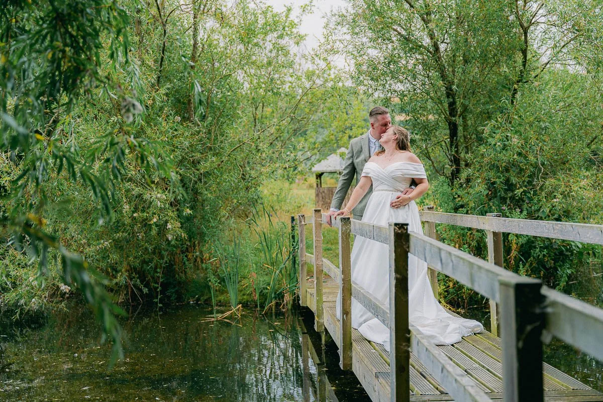 A couple dressed in wedding attire sharing a kiss on a small wooden bridge over a pond, surrounded by lush greenery dorset devon cinematic documentary style wedding photographer