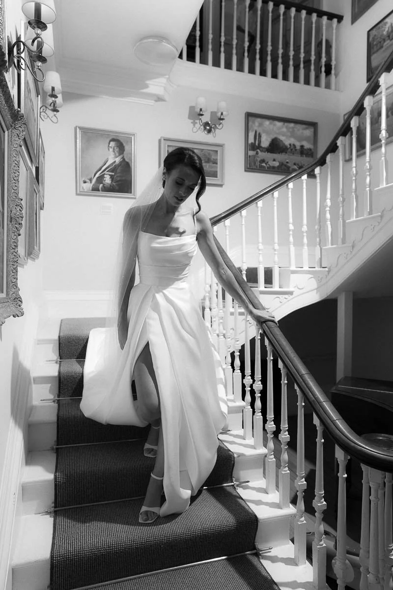 A woman in a wedding dress descending a staircase inside a house with framed artwork on the wall dorset devon cinematic documentary style wedding photographer