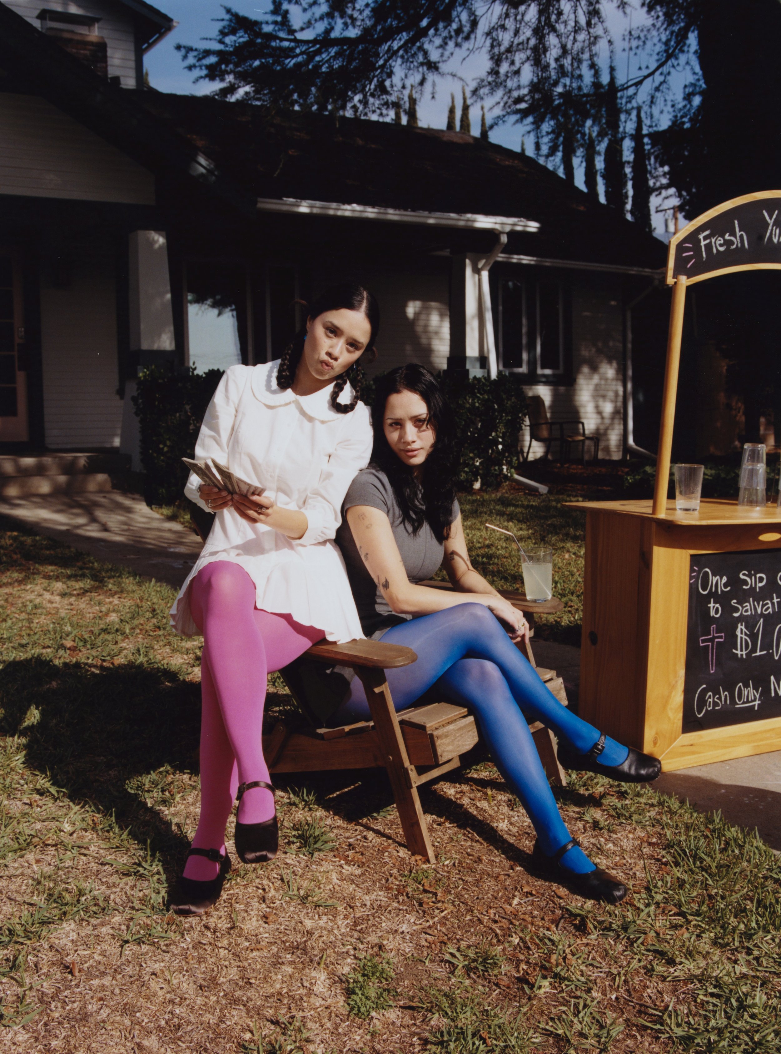Two young women with dark hair and dark lipstick sitting outside near a food stand that offers fresh drinks. One woman is wearing a white dress and pink tights, holding cash, and the other is in a gray T-shirt and blue tights. The setting is a yard w