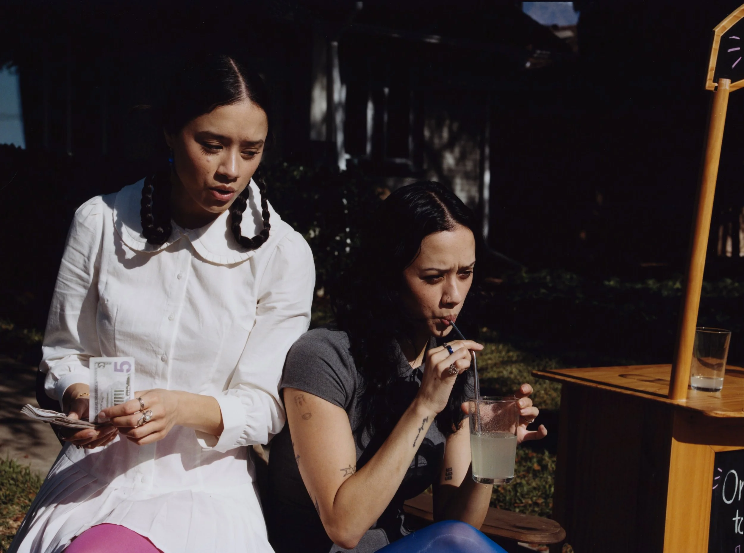 Two women are outdoors; one is holding cash and the other is drinking a lemonade with a paper straw.