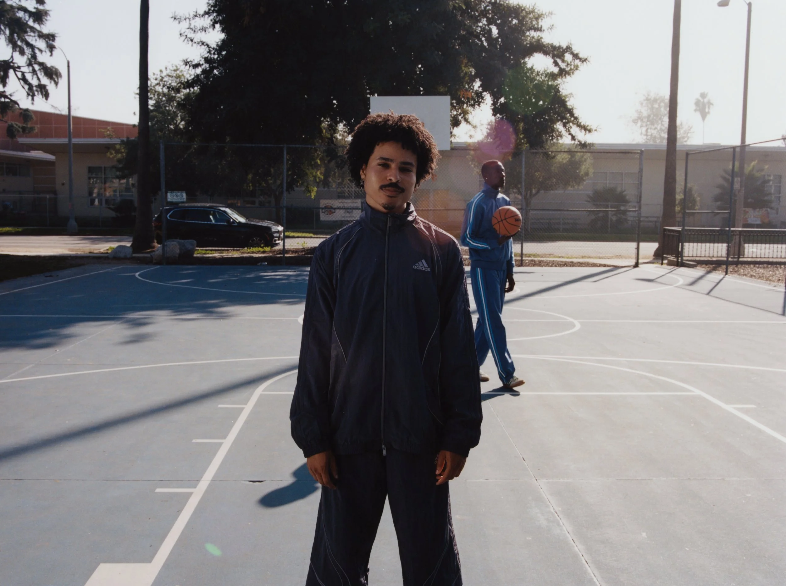 A young man with curly hair and a mustache wears a black Adidas tracksuit and stands on an outdoor basketball court. In the background, another person in a blue sports outfit holds a basketball.