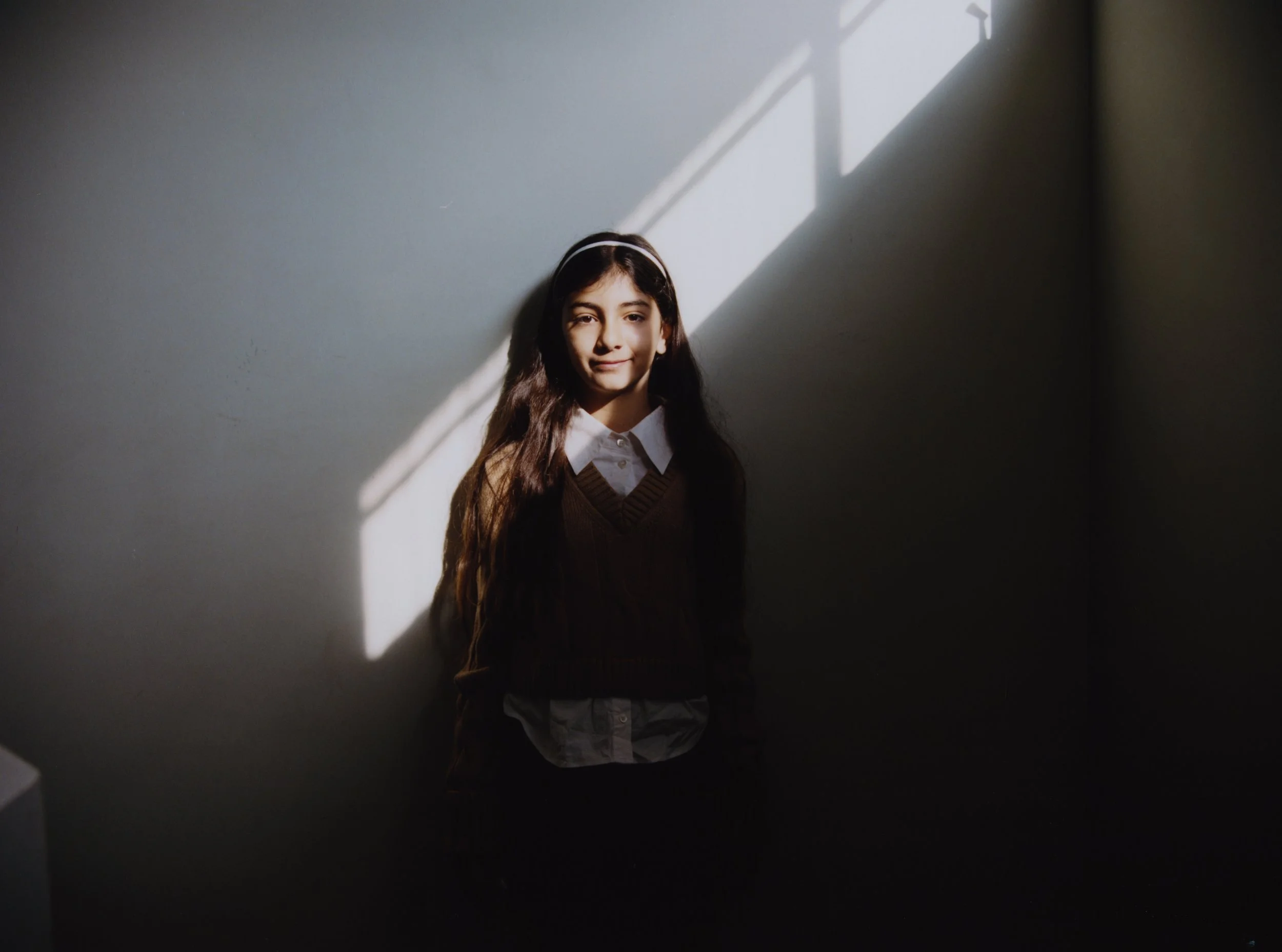 A young girl stands against a plain wall illuminated by diagonal sunlight from a window, casting a bright patch on the wall behind her.