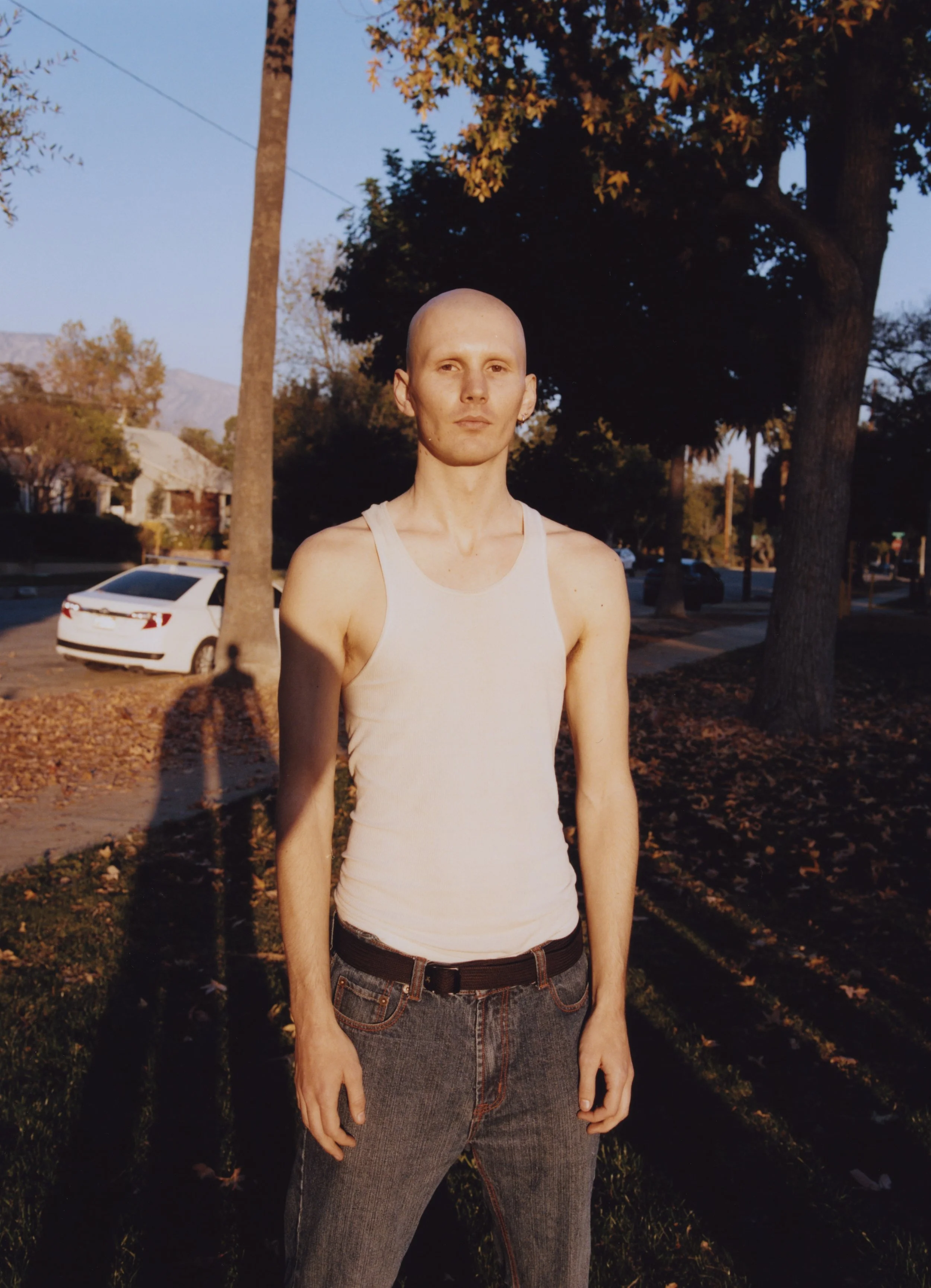 A young man with a shaved head wearing a white tank top and black jeans posing outdoors during late afternoon with trees and houses in the background.
