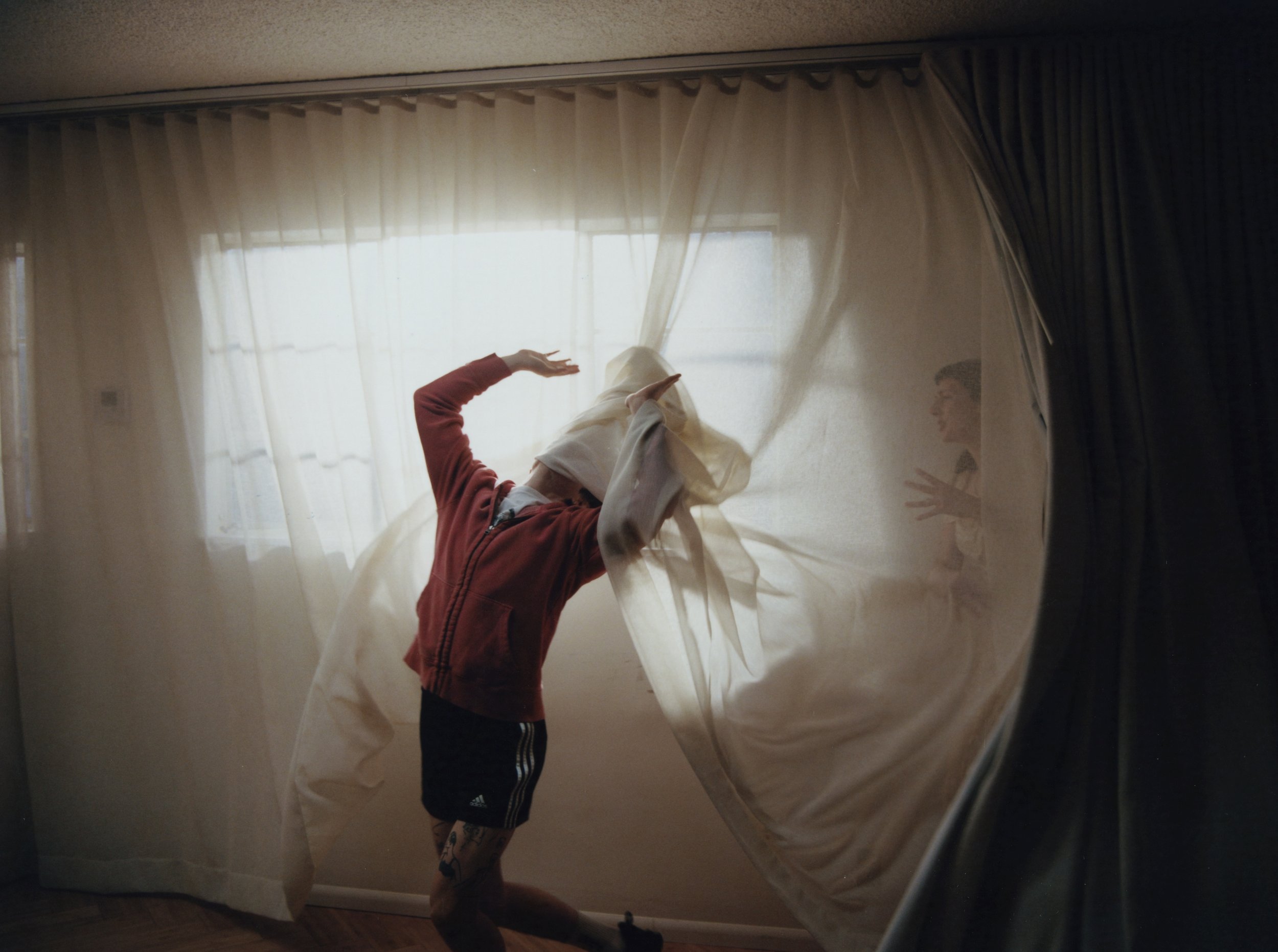 A person with a red hoodie, white face covering, and black shorts is playing behind a sheer curtain, with their arm raised and head turned to the side. A child is seen in the background through the curtain, laughing and reaching out.