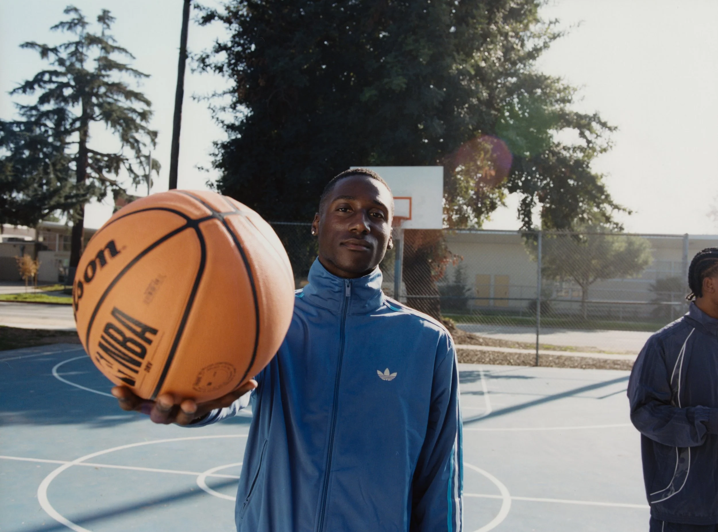 A young man holding a basketball on an outdoor court with trees and a basketball hoop in the background.