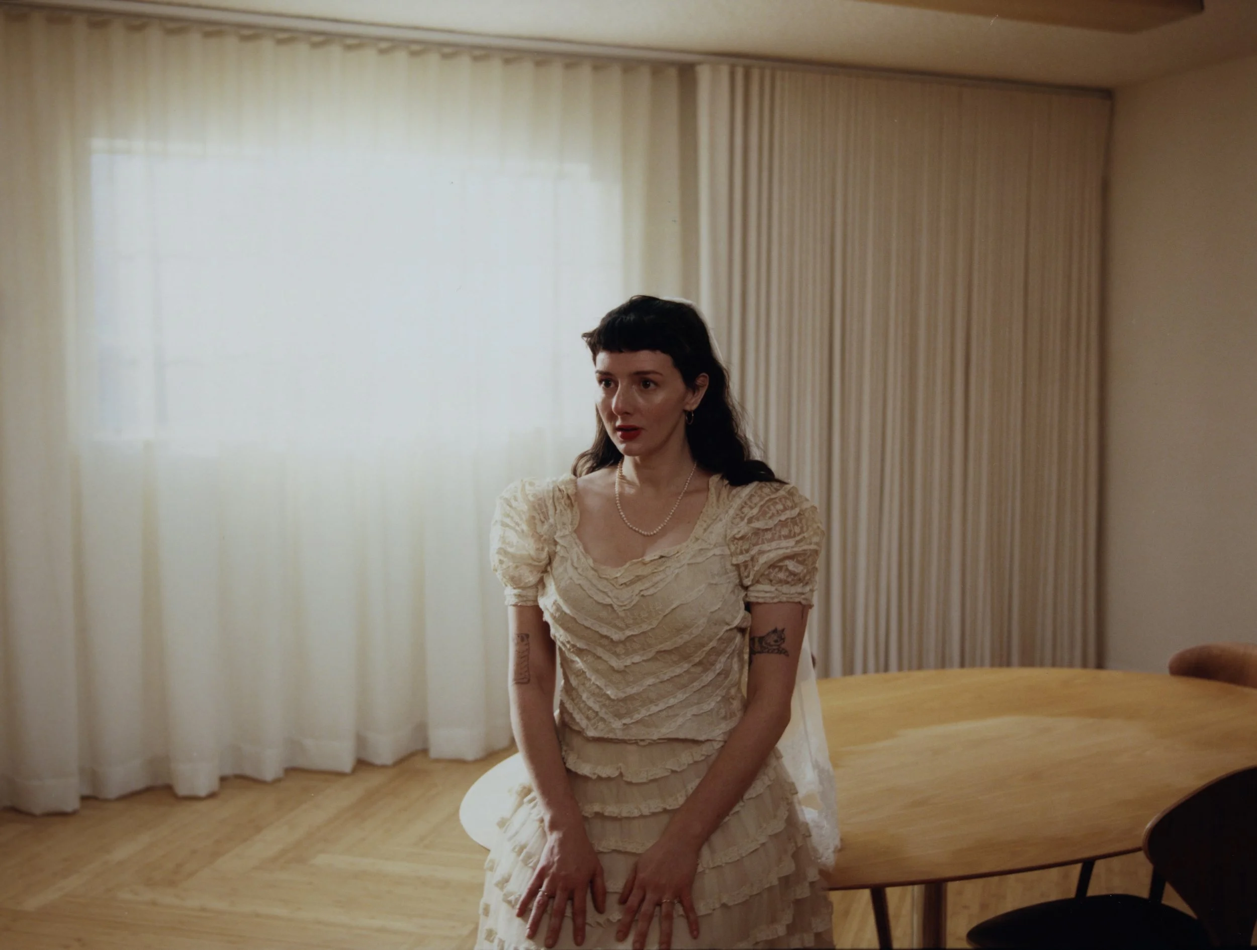 A woman with dark hair wearing a vintage style cream-colored dress with puffed sleeves and layered skirt, standing in a room with cream curtains and a wooden table.