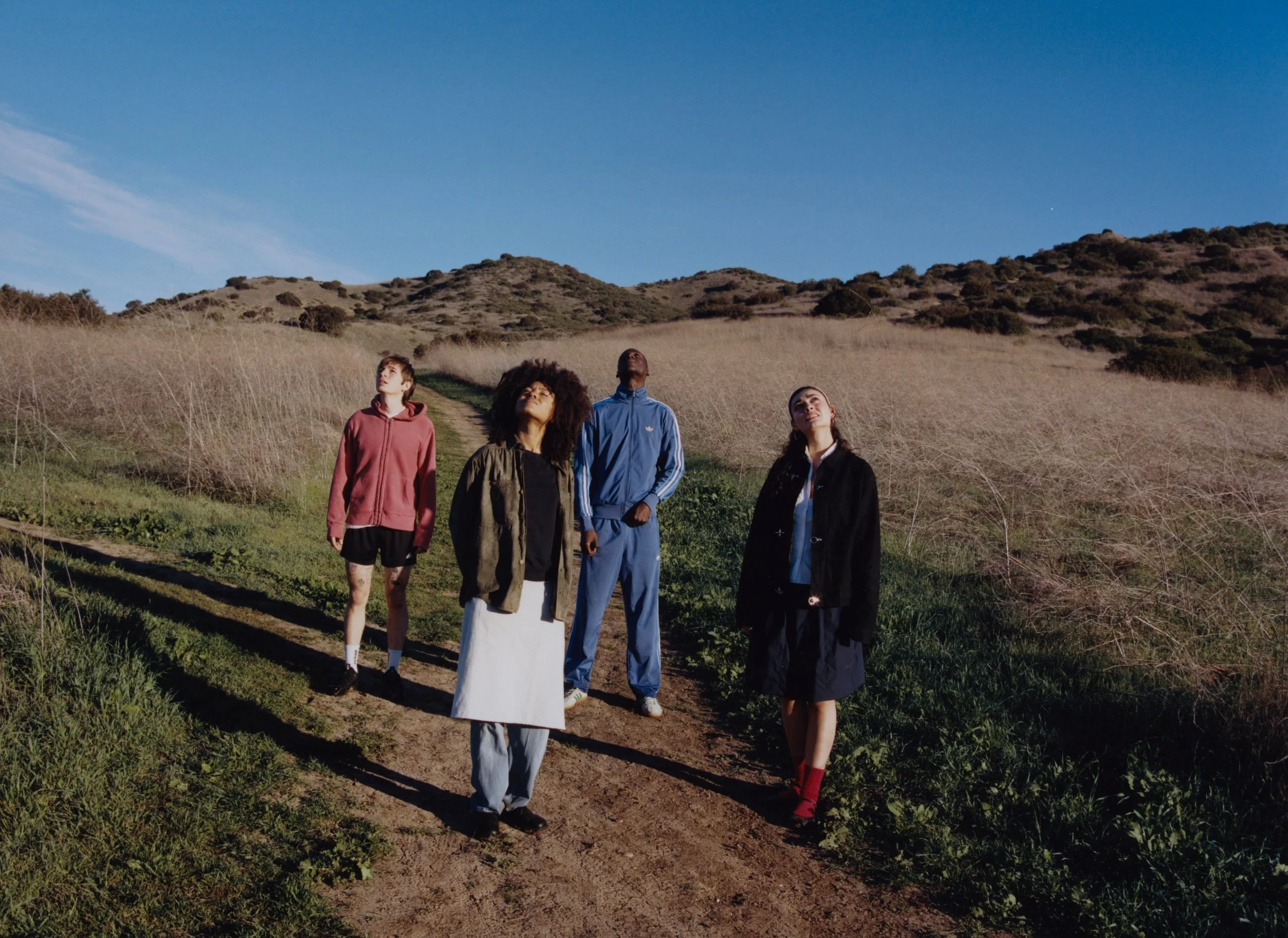 Four people stand on a dirt trail in a hilly outdoor landscape, looking up at the sky on a clear day.