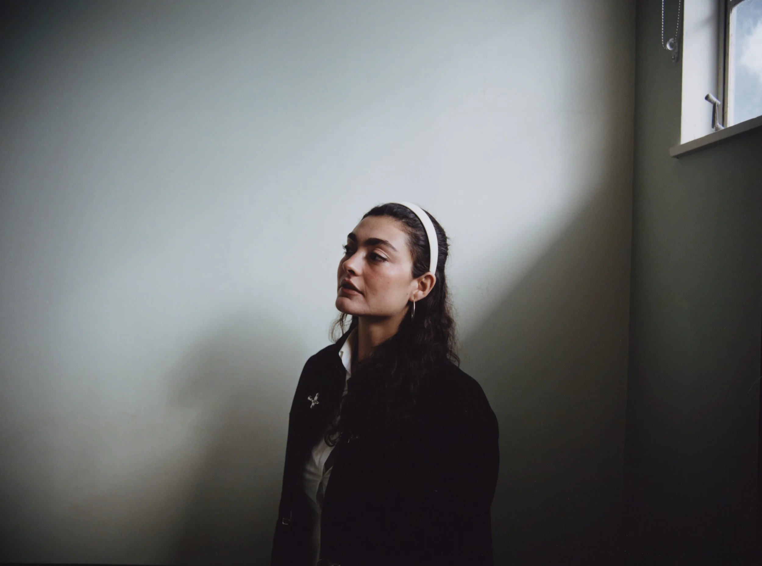 A woman with dark curly hair, wearing a white headband and a black jacket, standing in front of a white wall next to a window letting in natural light.
