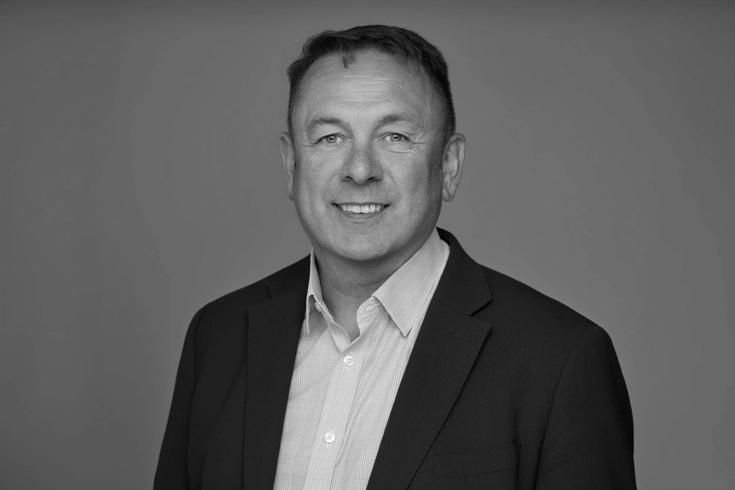 Black and white professional portrait of a man smiling, wearing a suit jacket and collared shirt, against a plain background.