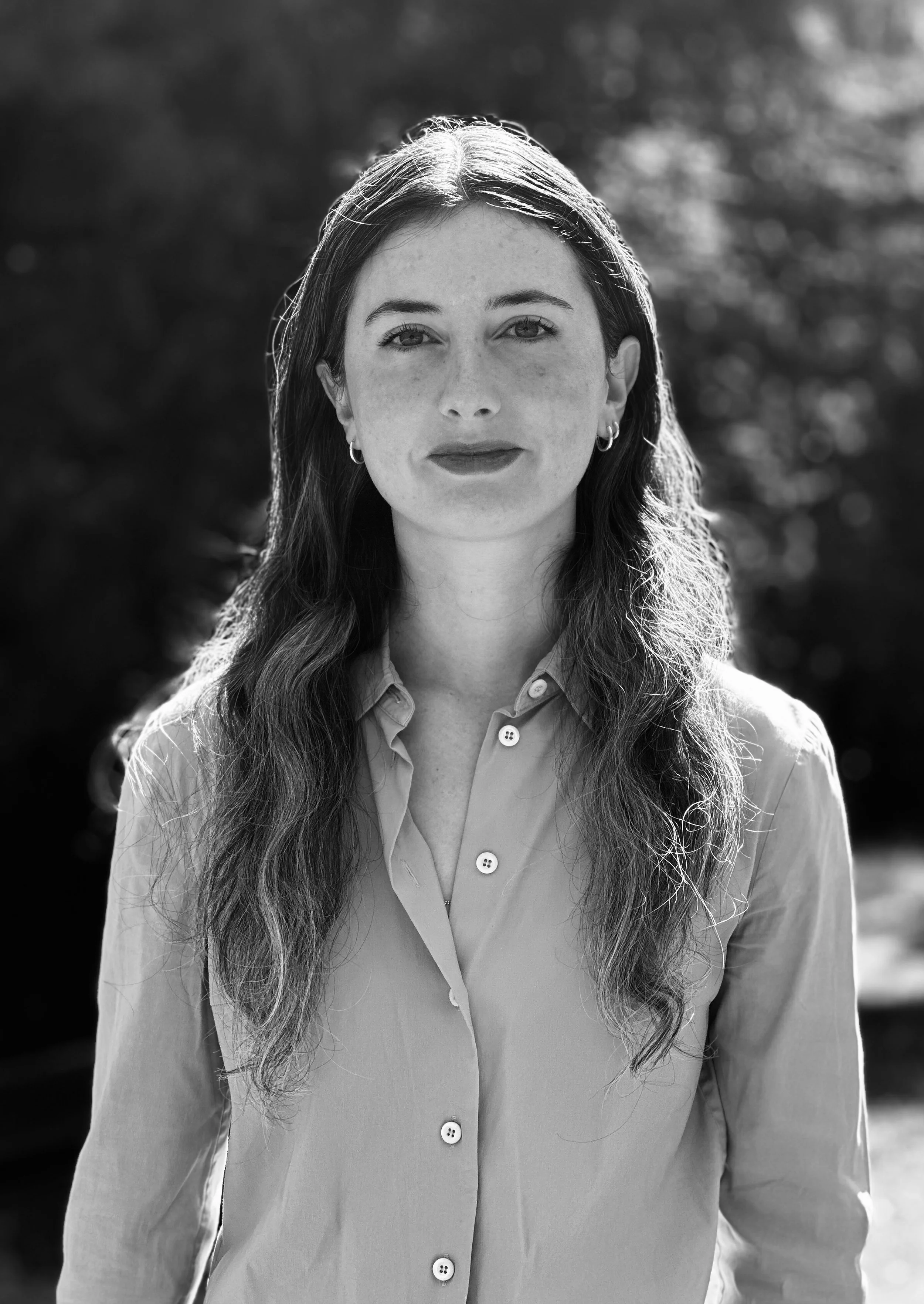 Black and white portrait of a woman with wavy hair, wearing earrings and a button-up shirt, outdoors with blurred trees in the background.